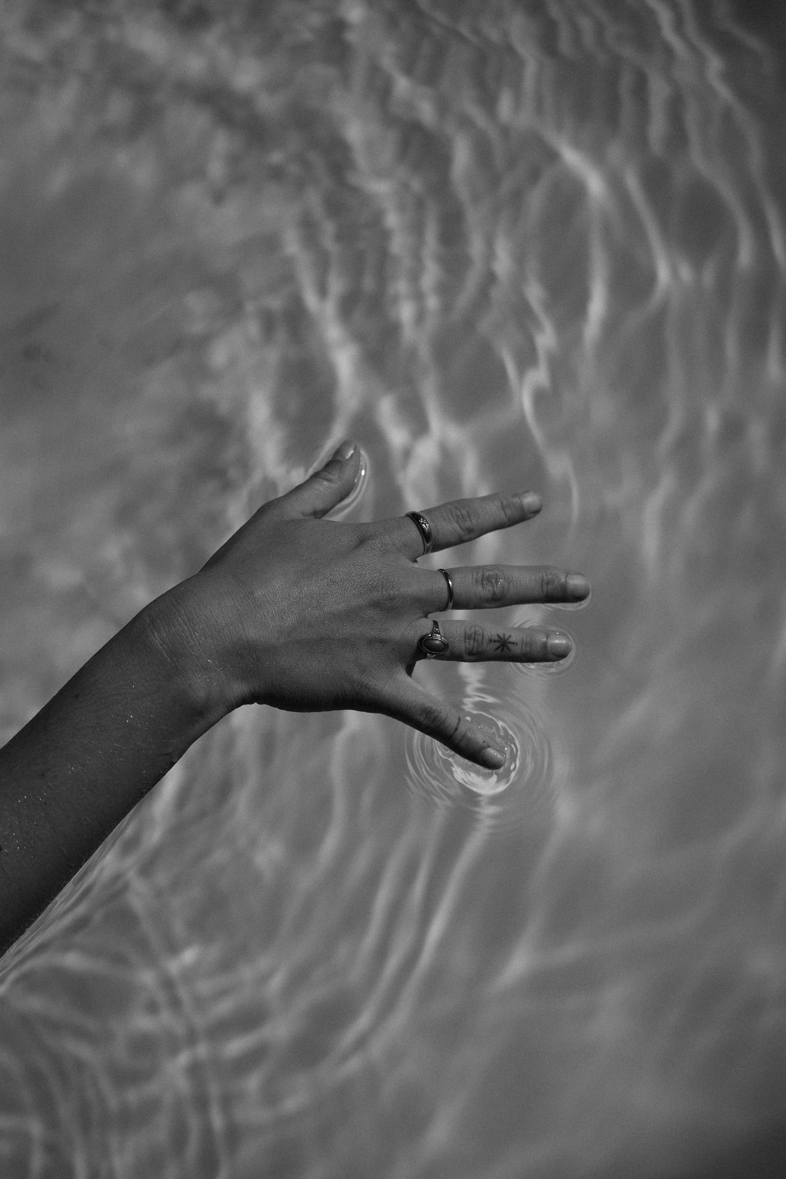 A black and white photo shows a hand with rings extended over the water, creating ripples that reflect light, suggesting a calm pool or ocean surface.