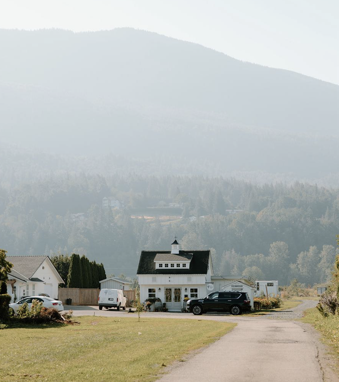 A small white house with a black roof in a rural area, surrounded by greenery and parked cars, with distant trees and mountains in the background.