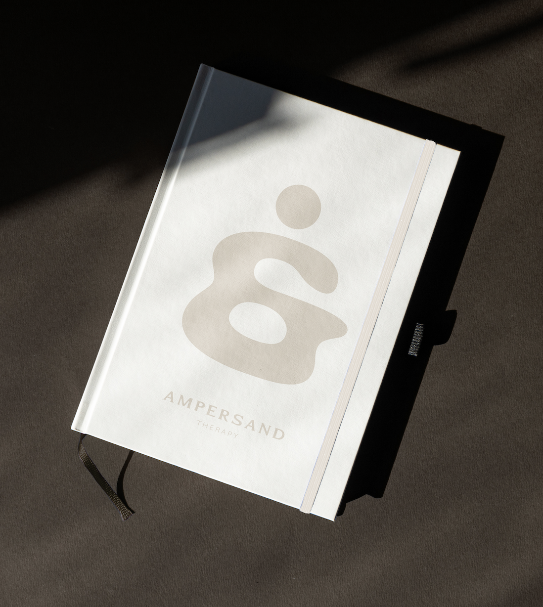 A white hardcover book titled 'Ampersand Therapy' with a minimalist ampersand symbol on the cover, lying on a brown surface with a light shadow.