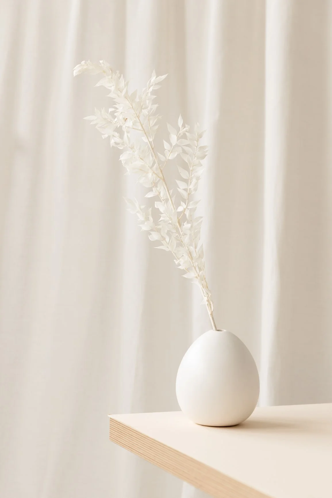 A white minimalist ceramic vase with a single branch of white leaves on a light wooden shelf, in front of a cream-colored curtain.