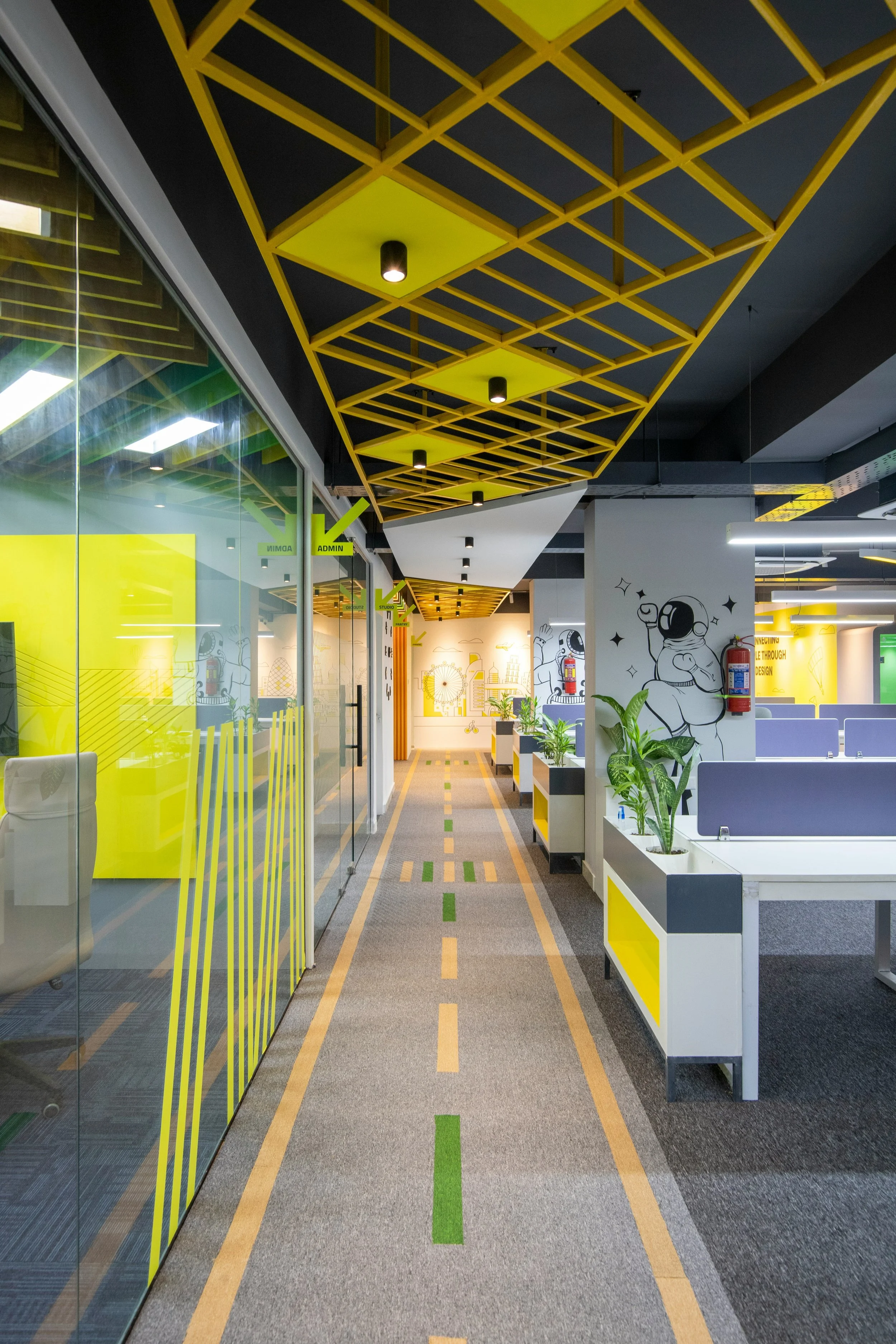 Modern office corridor with yellow geometric ceiling design, glass walls on the left, white desks with plants on the right, and travel-themed wall art. Signage indicates 'Admin' and 'Studio' areas.
