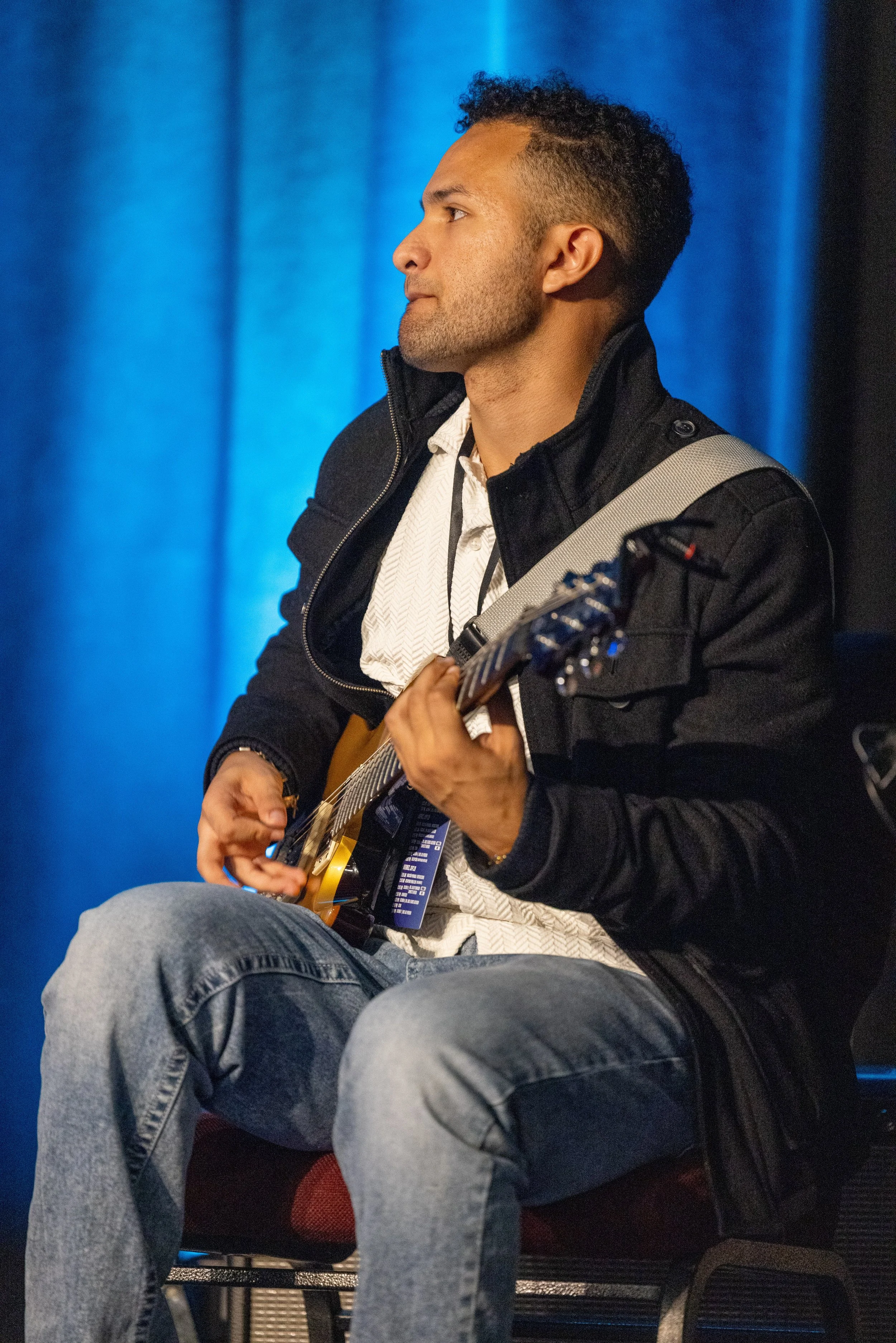 A young man with curly hair and a fade haircut, sitting on a chair, playing an acoustic guitar, wearing a black jacket over a white shirt, against a blue curtain background.