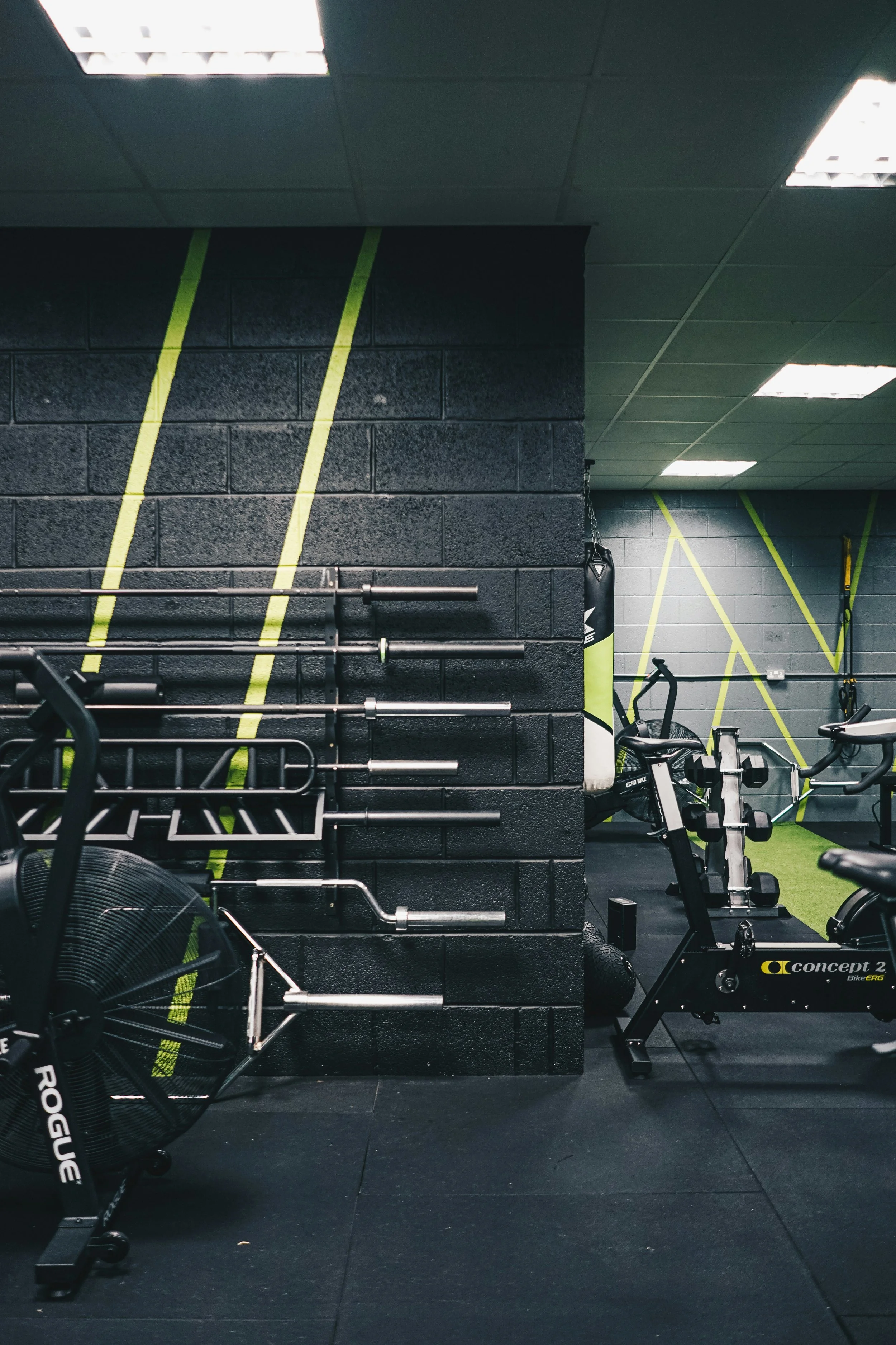 Interior of a gym with workout equipment including rowing machines, stationary bike, weights, and black brick walls with green diagonal lines.