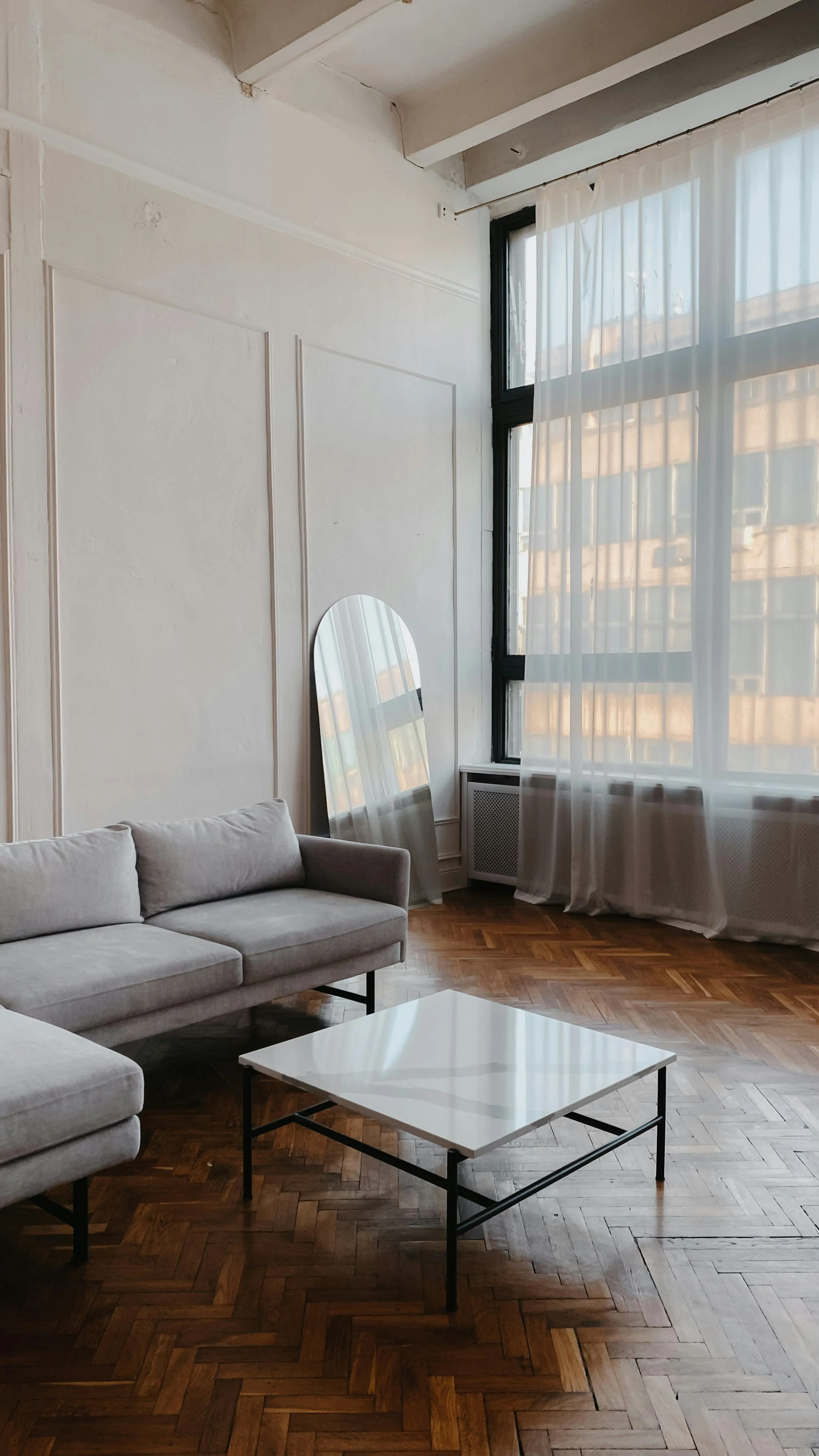 A living room with a gray sofa, a white marble coffee table, a tall mirror leaning against the wall, large windows with sheer white curtains, and hardwood parquet flooring.