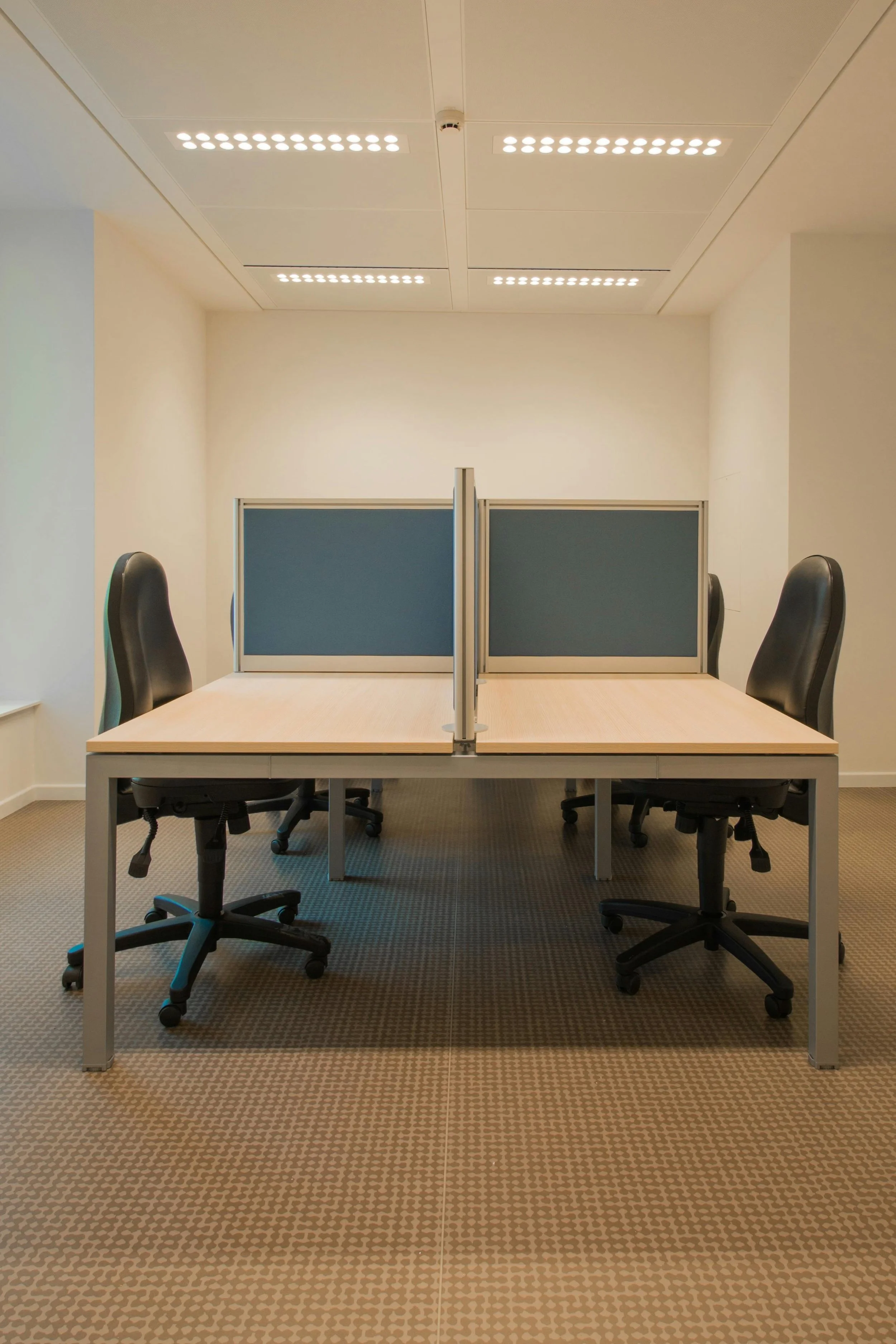 Empty office space with two desks separated by a divider and four chairs, two on each side. There are grid-patterned carpet and white walls with ceiling lights.