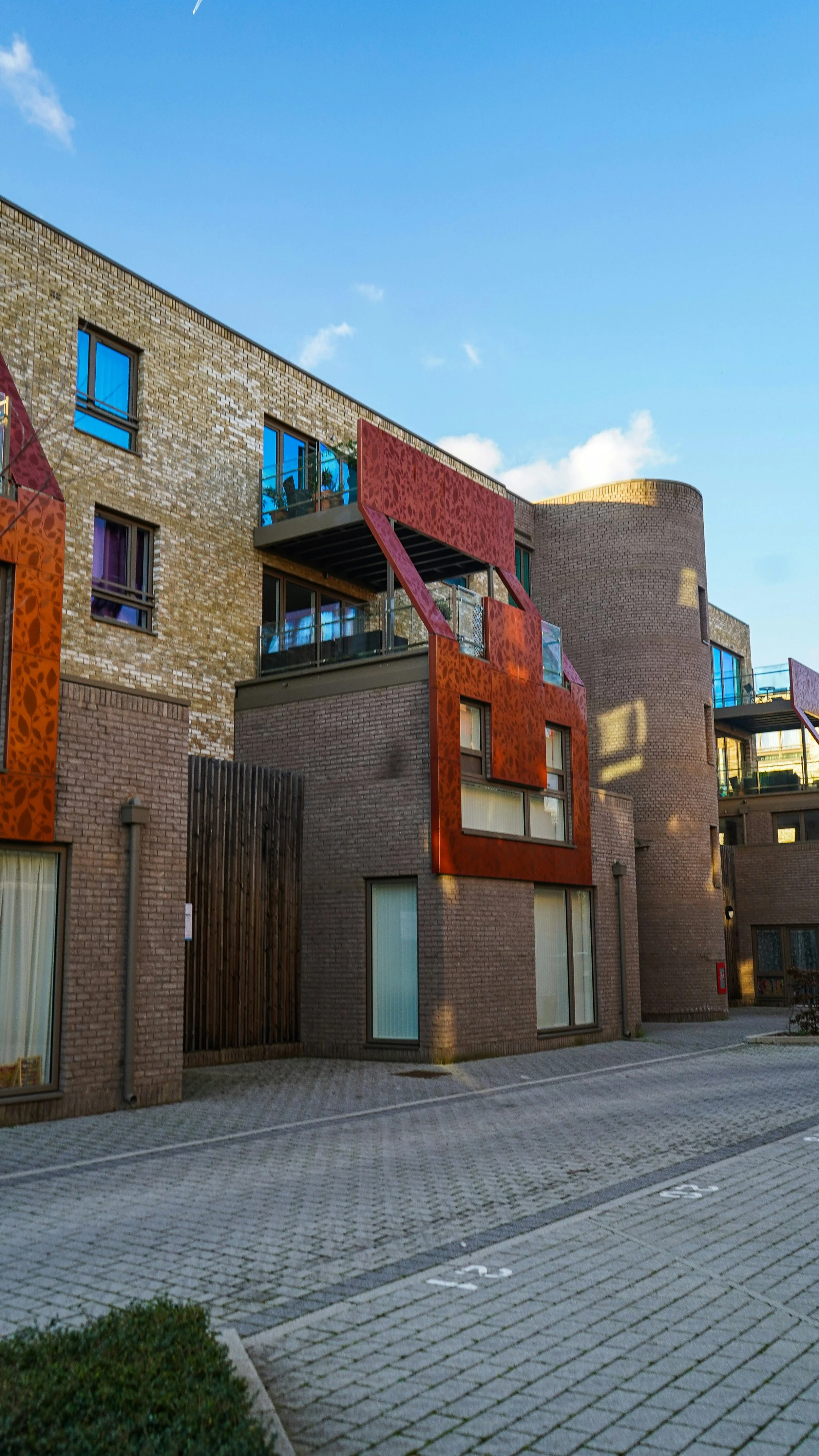 A modern apartment building with a brick facade and colorful decorative panels, with a parking lot in the foreground and a partly cloudy sky above.