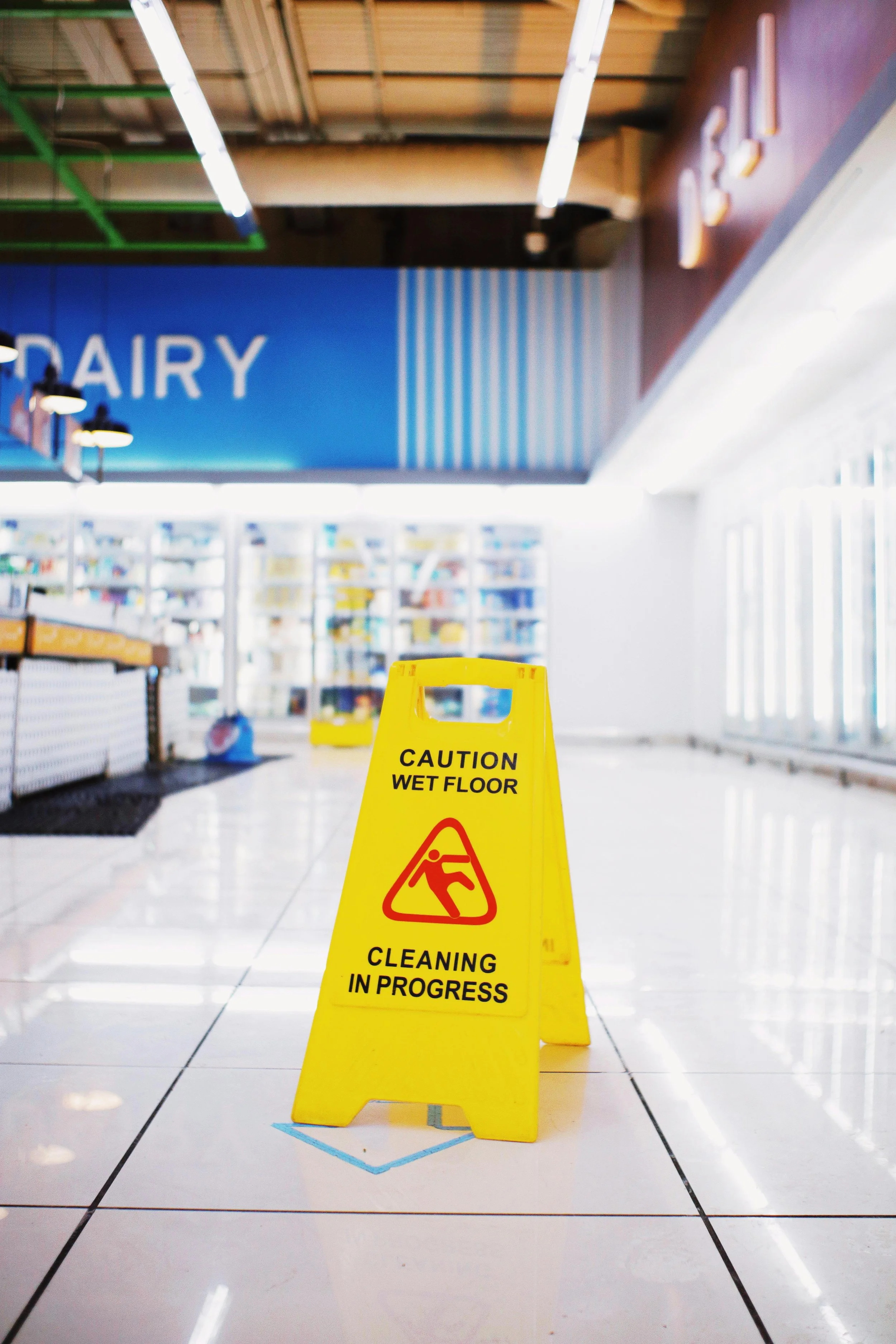 A yellow caution sign on tiled floor indicating wet floor and ongoing cleaning inside a store or mall, with shelves and a large blue 'Dairy' sign in the background.