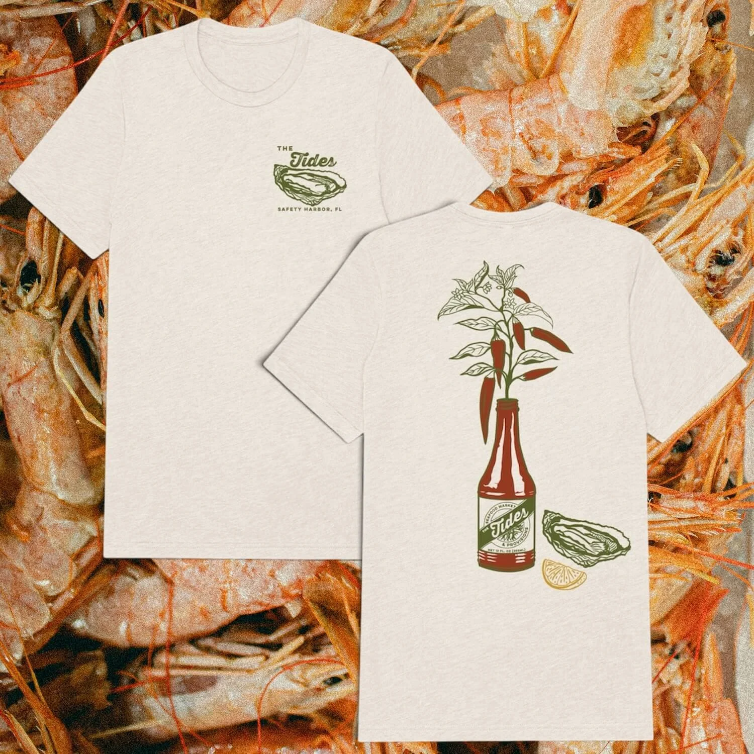 Two white T-shirts with vintage-style designs, placed on a background of shrimp and shells. The front shirt features a green and red illustration of a bottle with a plant growing from it, and a lemon wedge at the bottom. The back shirt has the text "The Tides" with a drawing of a shell, and "Safety Harbor, FL" underneath.