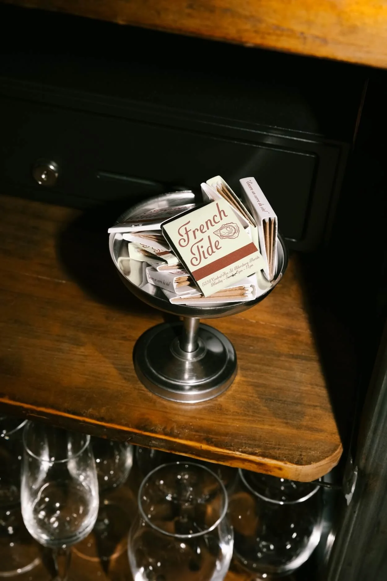 A cocktail stirrer glass filled with matchbooks labeled 'French Tide' on a wooden shelf with wine glasses below.