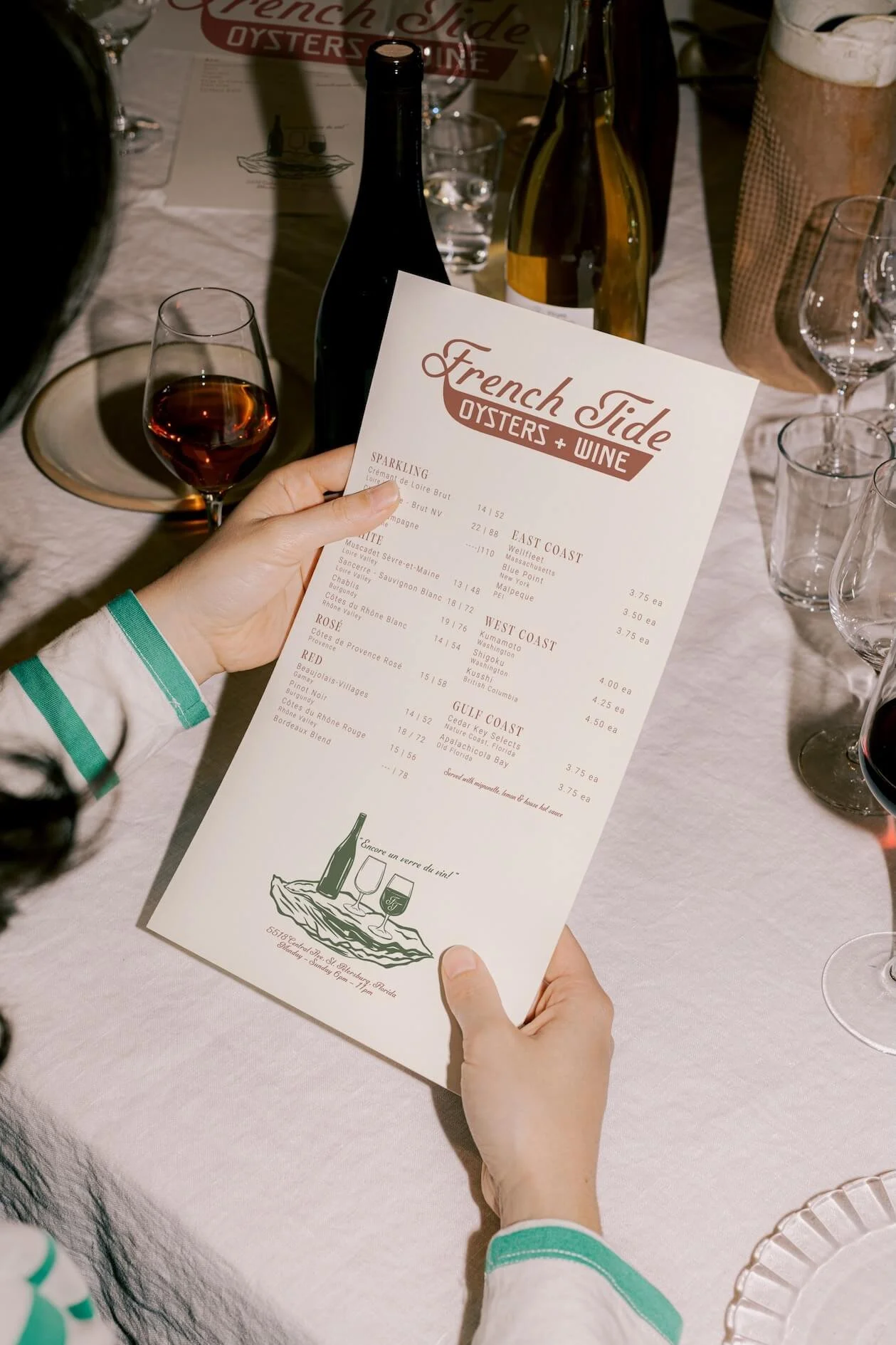 A person holding a wine menu from French Tide Oysters + Wine restaurant, with wine glasses and bottles on a table.