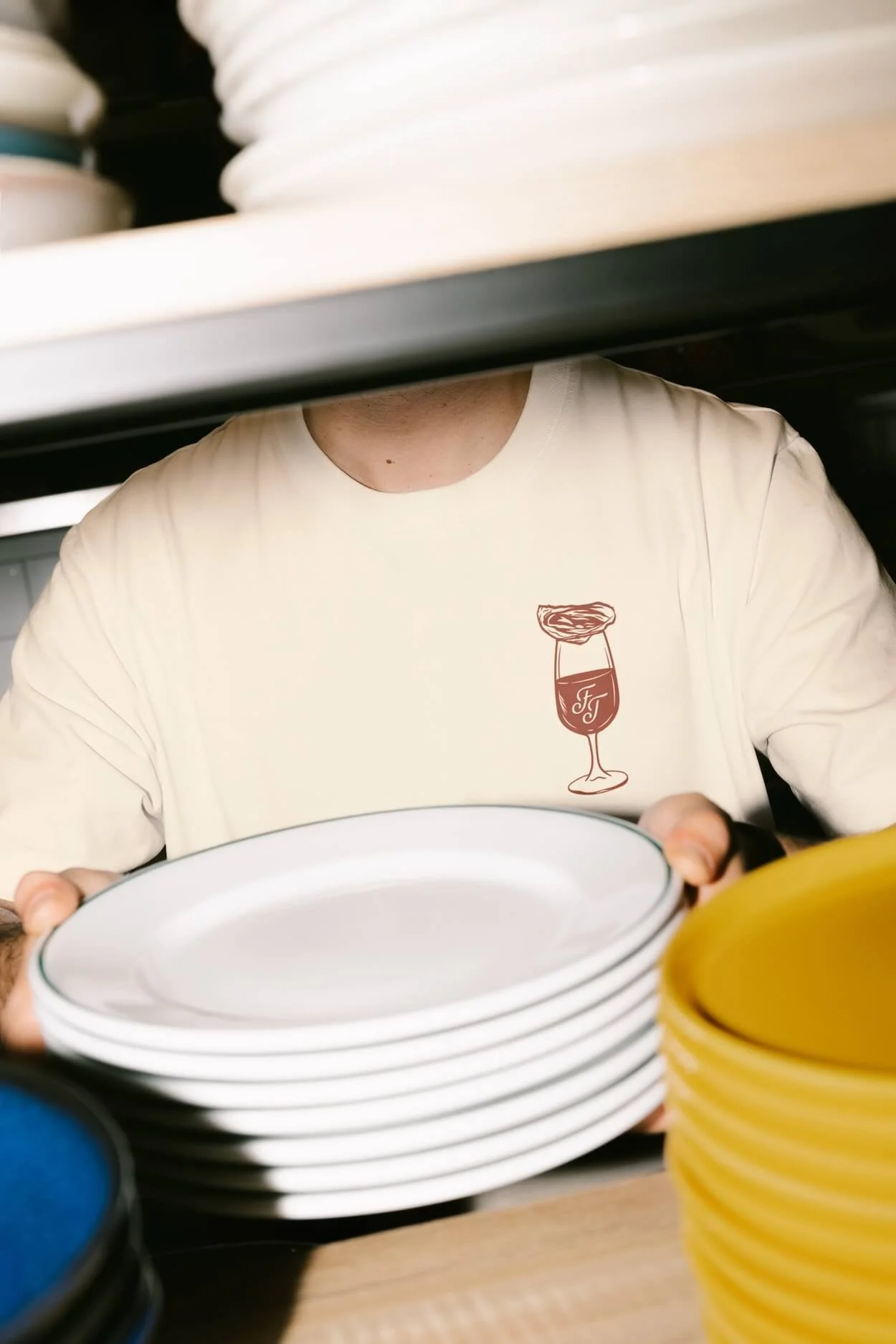 Person holding white plates in a kitchen or restaurant setting, seen partially with shelves of dishes above and colorful bowls nearby.