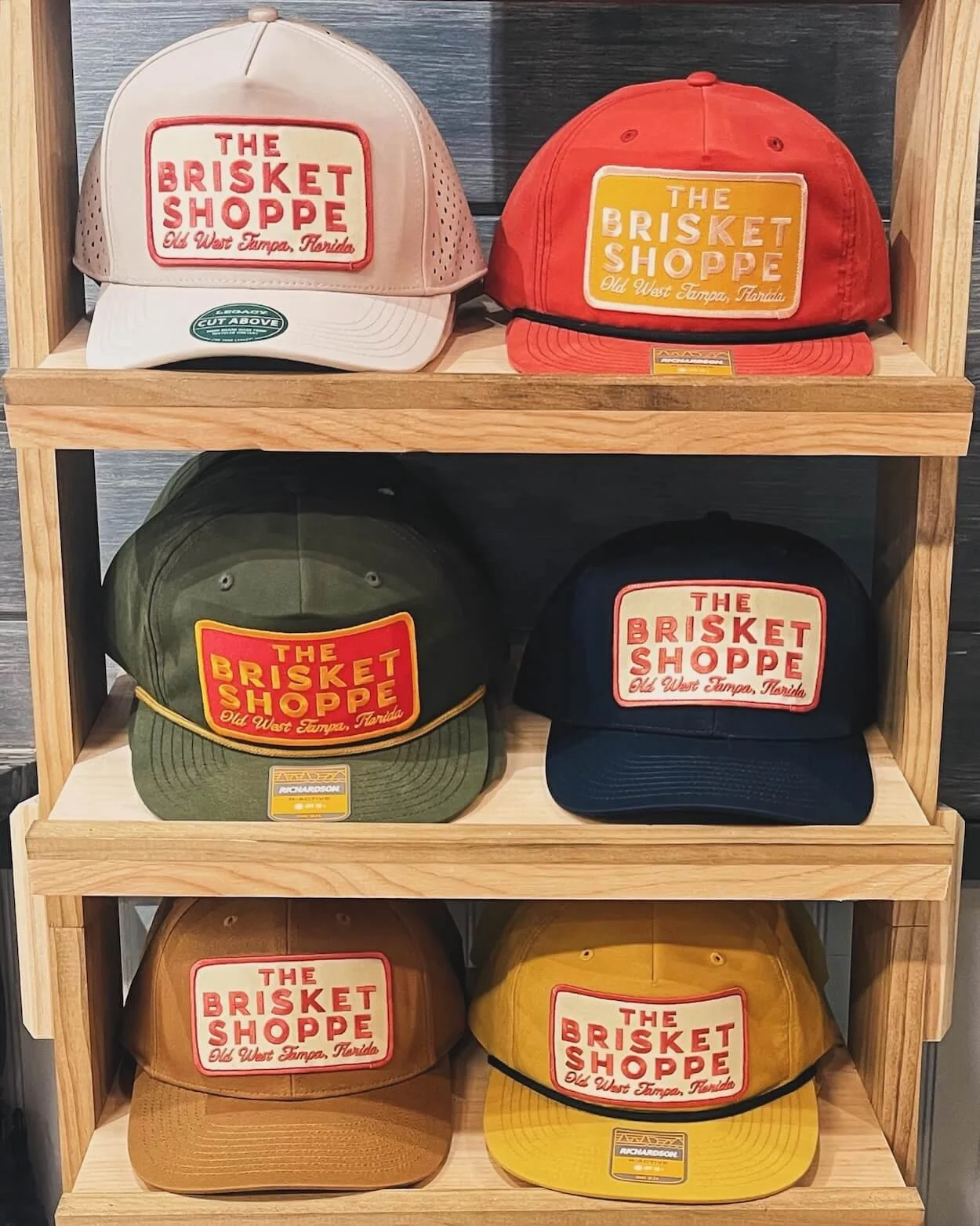 Collection of six caps with embroidered patches saying "The Brisket Shoppe, Old West Tampa, Florida," in beige, orange, green, navy, tan, and yellow, displayed on a wooden shelf.