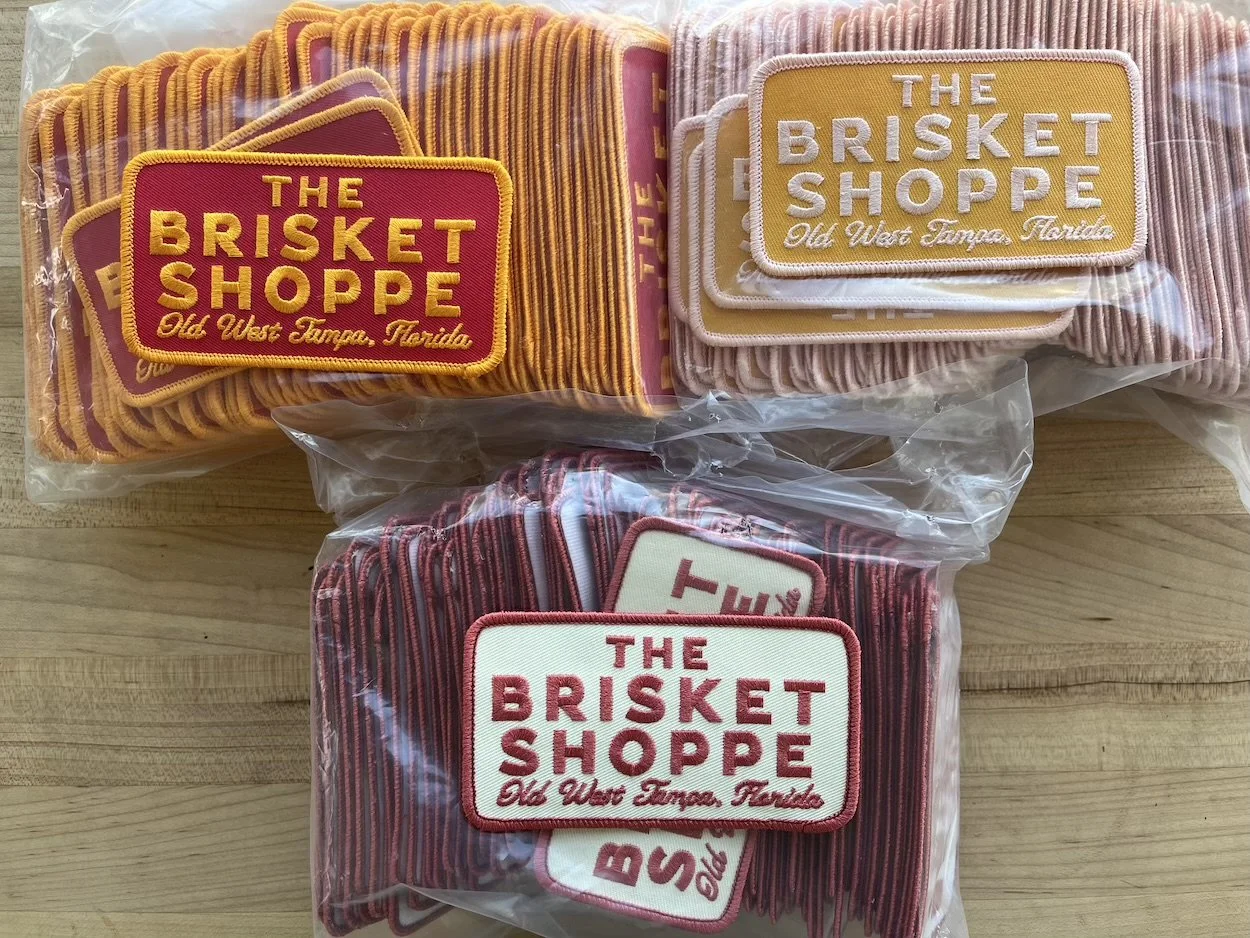 Three packs of fabric patches with embroidered text for The Brisket Shoppe, Old West Tamps, Florida, in yellow, pink, and red color schemes, packaged in plastic bags and arranged on a wooden surface.
