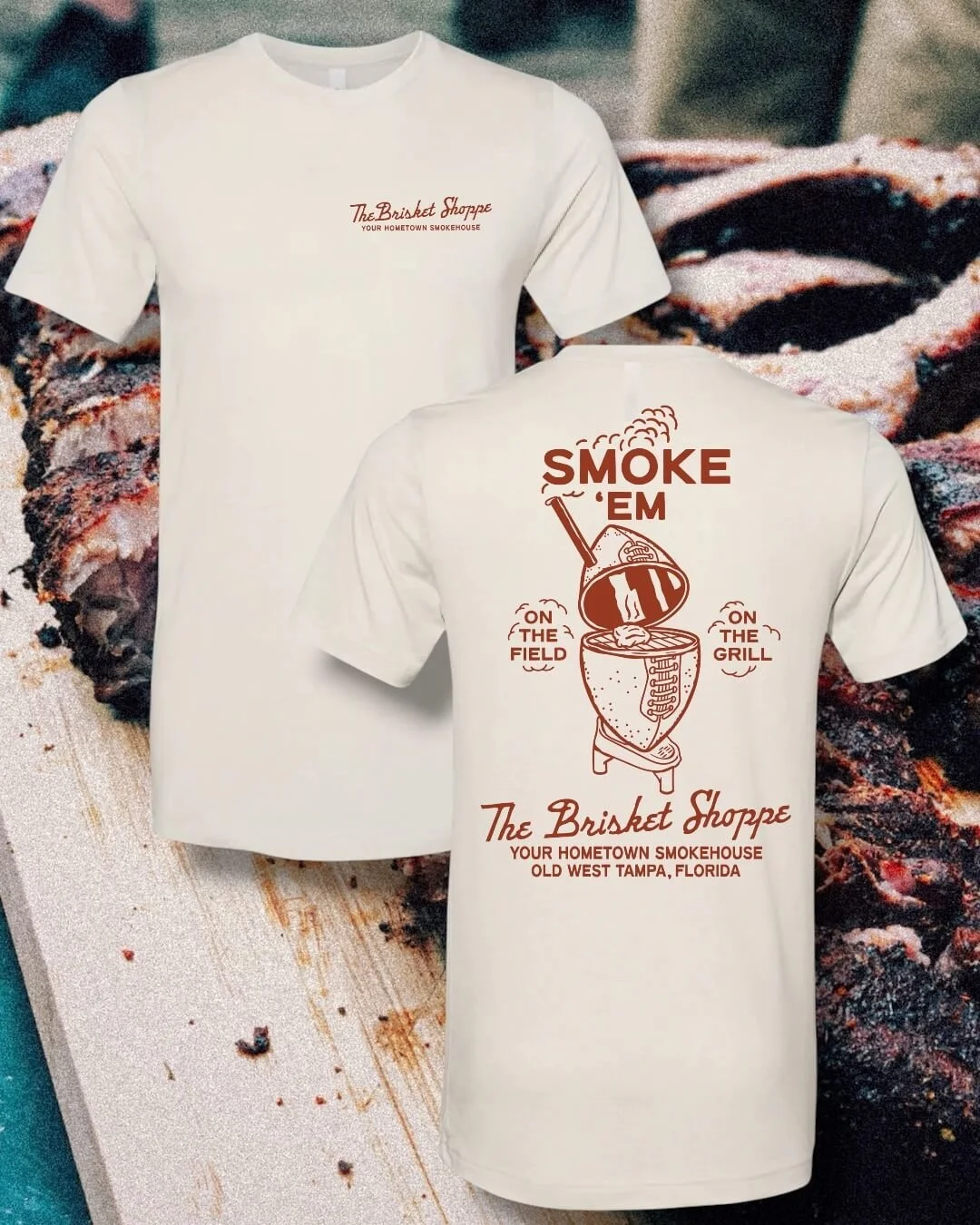 White t-shirt with a logo that reads 'The Brisket Shoppe, Your Hometown Smokehouse' on the front, and a graphic of a smoker with the text 'Smoke 'Em on the Field on the Grill' and 'The Brisket Shoppe, Old West Tampa Florida' on the back. Background features cooked barbecue meat.