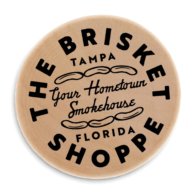A round wooden sign with black text that reads "The Brisket Shoppes" around the top and bottom, "Tampa Florida" in the middle, and "Your Hometown Smokehouse" in cursive in the center, with decorative swirls.
