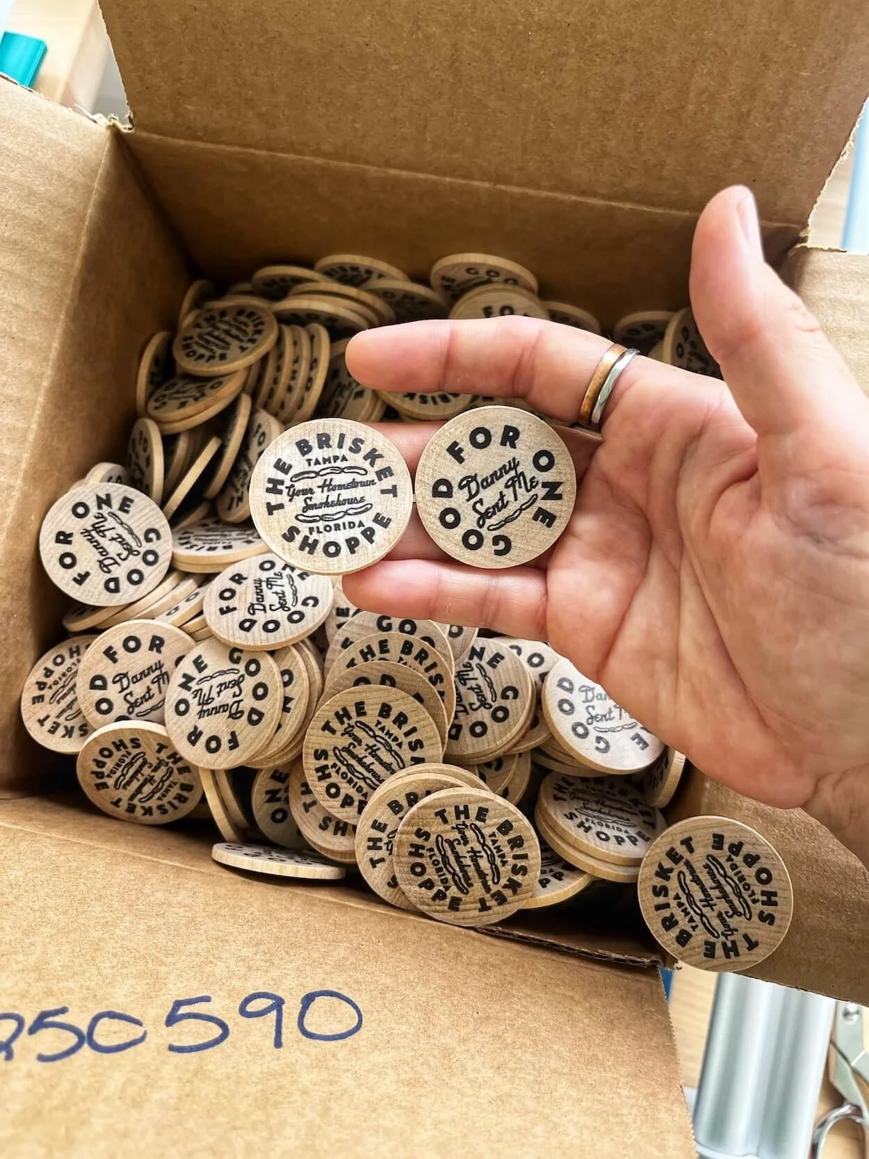 A person's hand holding a wooden token with text, surrounded by similar tokens in a cardboard box.
