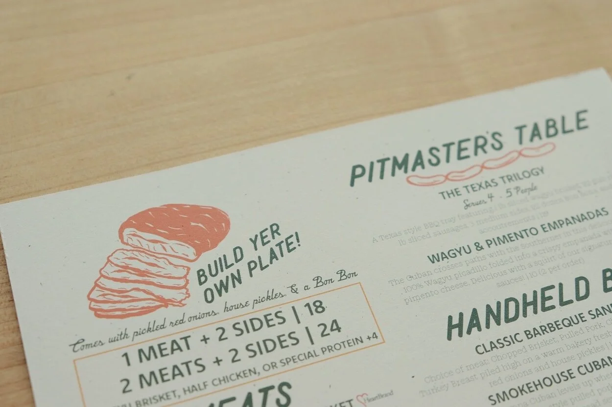 Part of a menu titled "Pitmaster's Table" featuring illustrations of sliced smoked meat with text promoting building your own plate with vegetables and sides, and highlighting options for meat with sides and protein.