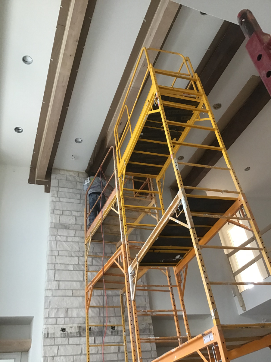Professional painters staining exposed ceiling beams in luxury home interior using scaffolding for detailed high-ceiling wood finishing and custom interior craftsmanship.