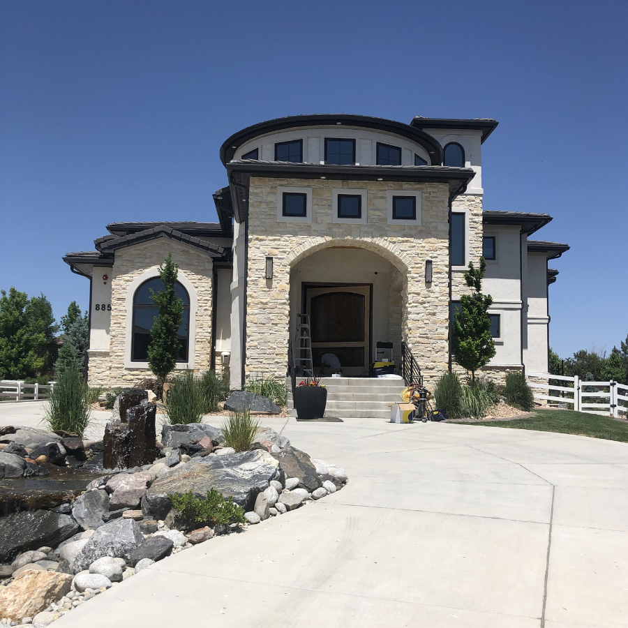 A large, modern house with a stone facade, multiple windows, and a round turret on the upper level. The front entrance has stairs leading up to a covered porch. The house is surrounded by landscaping, including trees, rocks, and a small pond with water features. The sky is clear and blue.