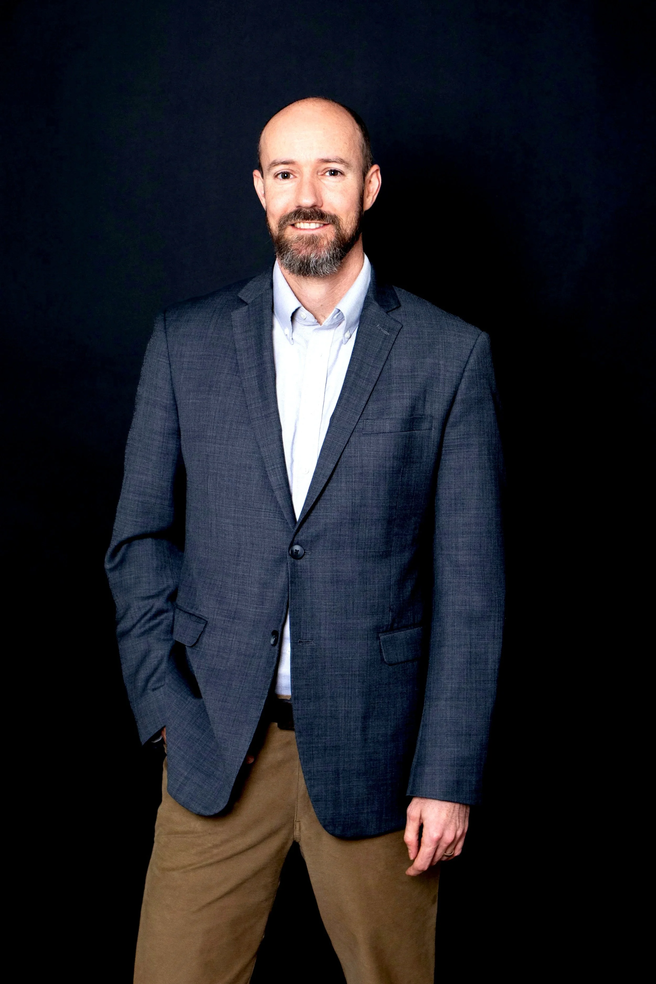 Dr. Andrew Rostenberg a navy blazer, light blue shirt, and tan pants standing against a black background.