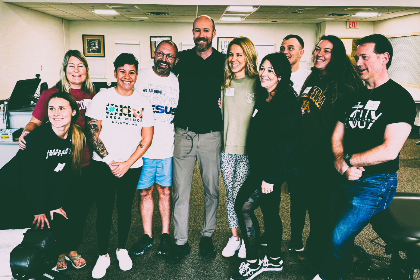 Group of ten diverse smiling people standing together indoors, some wearing casual clothes with logos or text, in a room with framed pictures on the wall.
