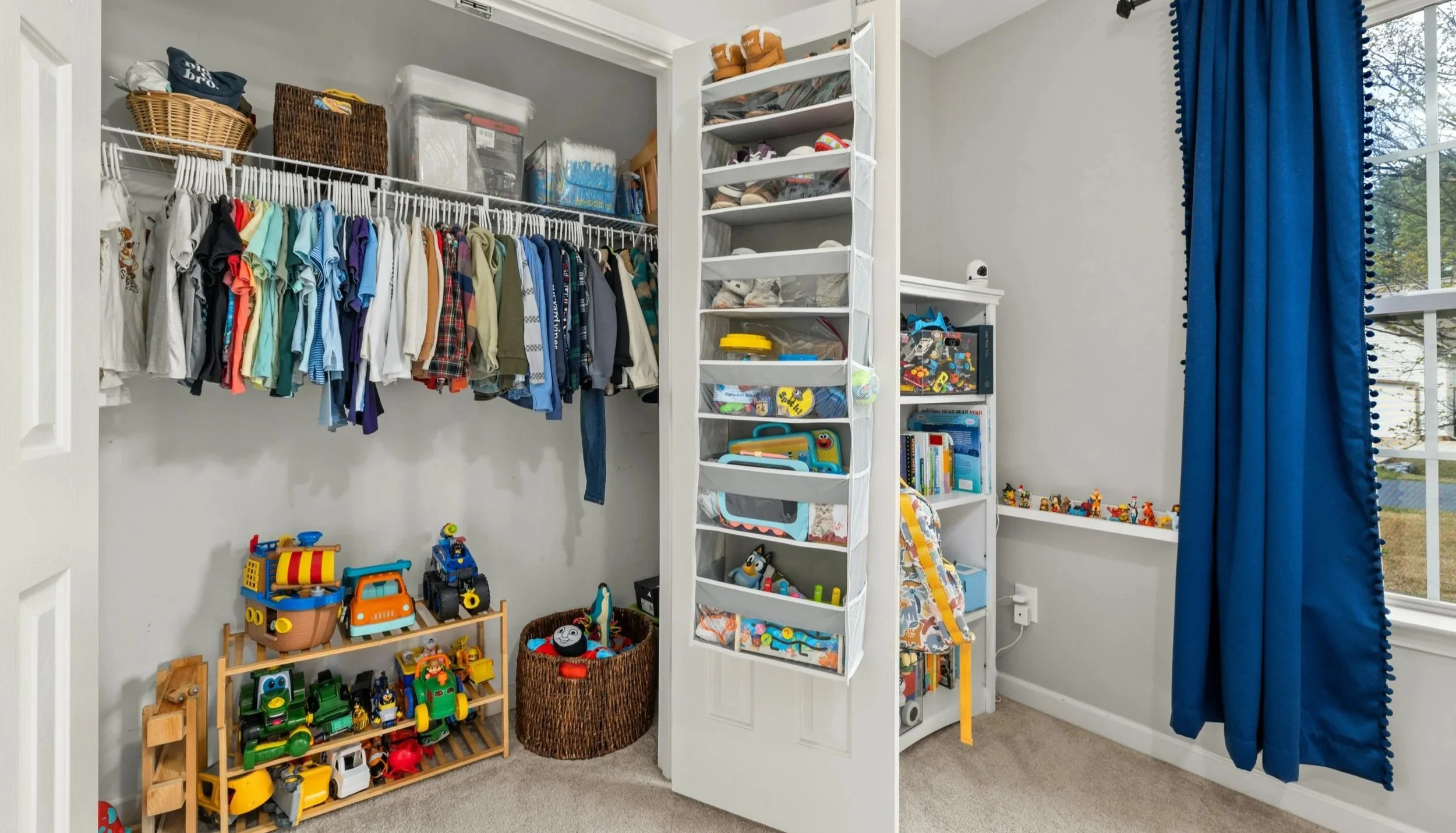 Child's closet with hanging clothes, toy shelves, and bins for toys and books.