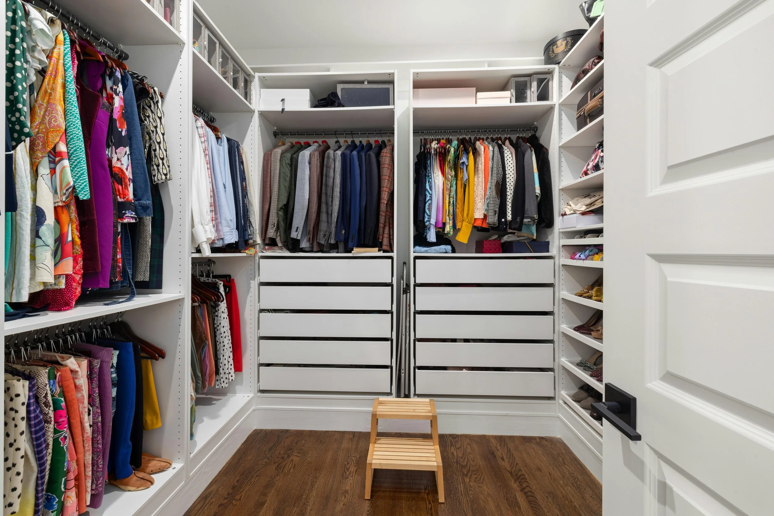 A walk-in closet with white shelves filled with clothes, shoes, and storage boxes, a small wooden stool in the center, and a wooden floor.