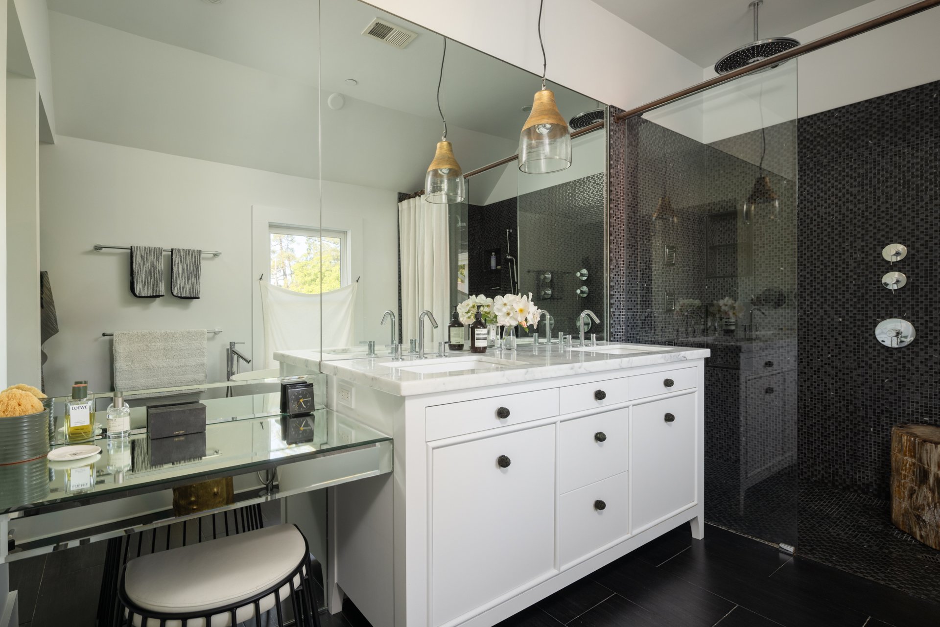 Modern bathroom with white vanity, marble countertop, black knobs, glass shower with black mosaic tiles, and large mirror. Decor includes flowers, toiletries, and hanging pendant lights.