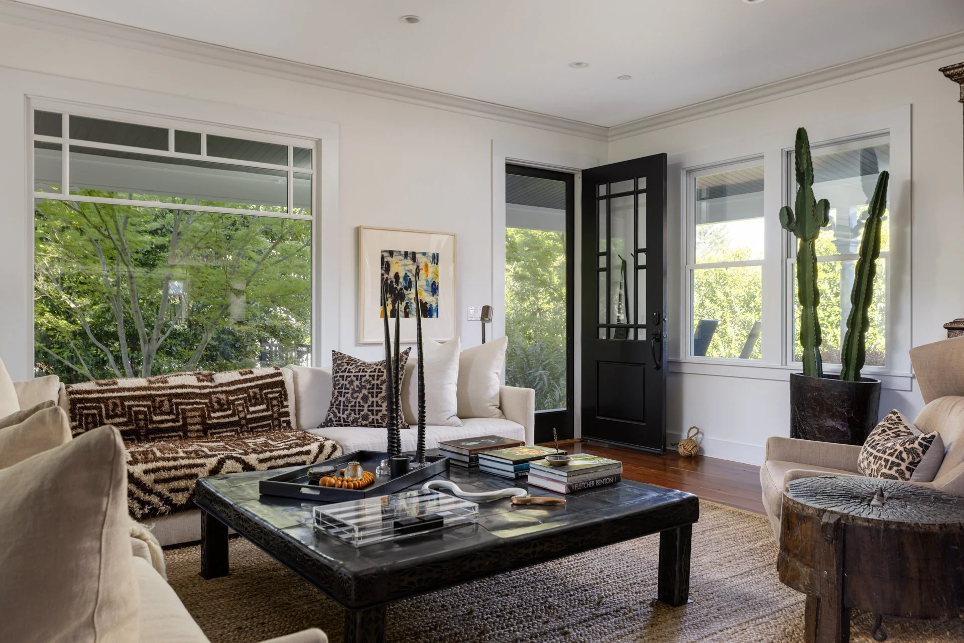 Living room with white walls, large windows showing green trees outside, a black front door, and furniture including a beige sofa, a black coffee table, and a large potted cactus plant.