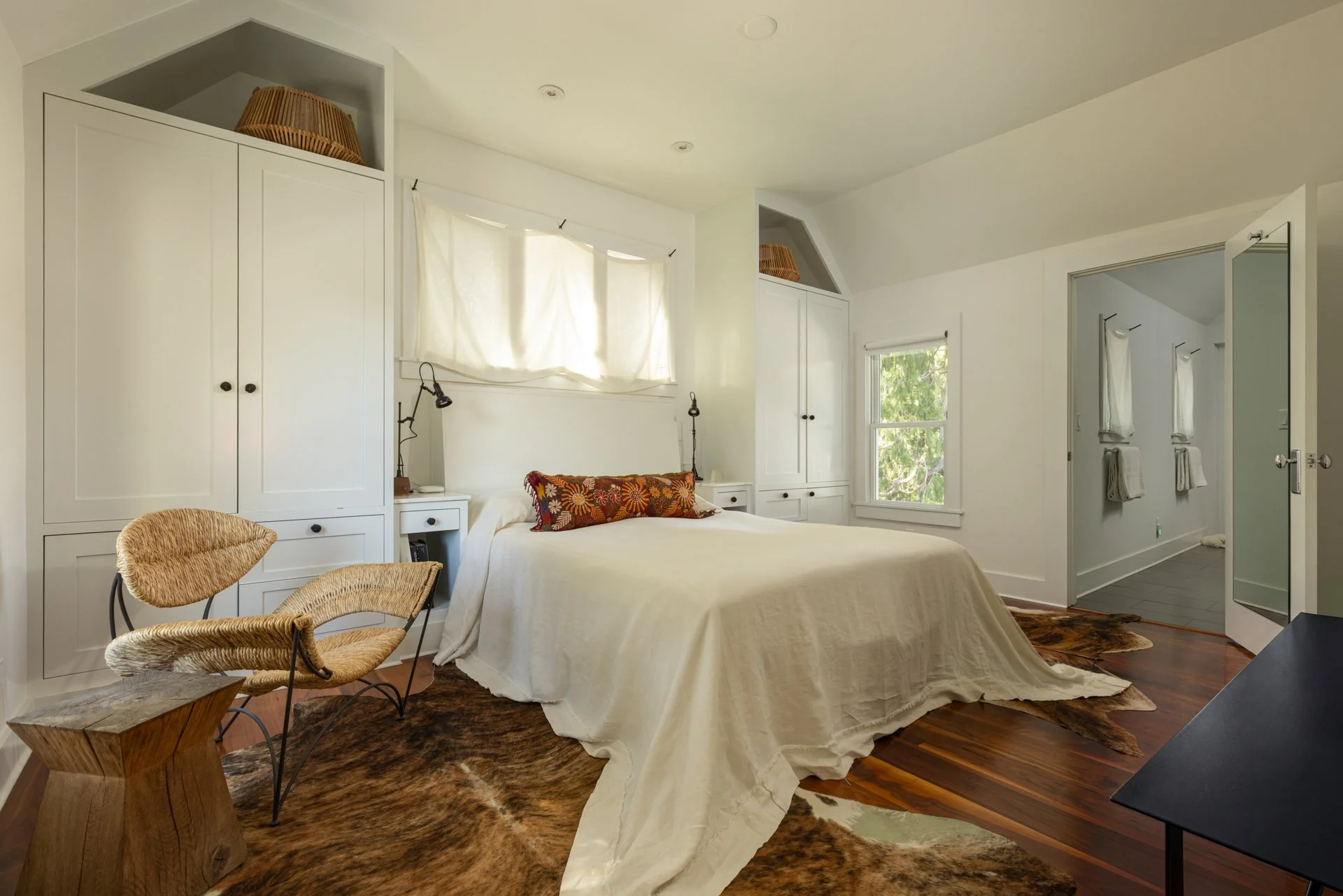 A cozy bedroom with a large bed covered in white bedding, decorated with a colorful floral pillow. To the left, a small woven chair with a curved backrest sits at a rustic wooden side table placed on a cowhide rug. Behind the bed, there are white bui