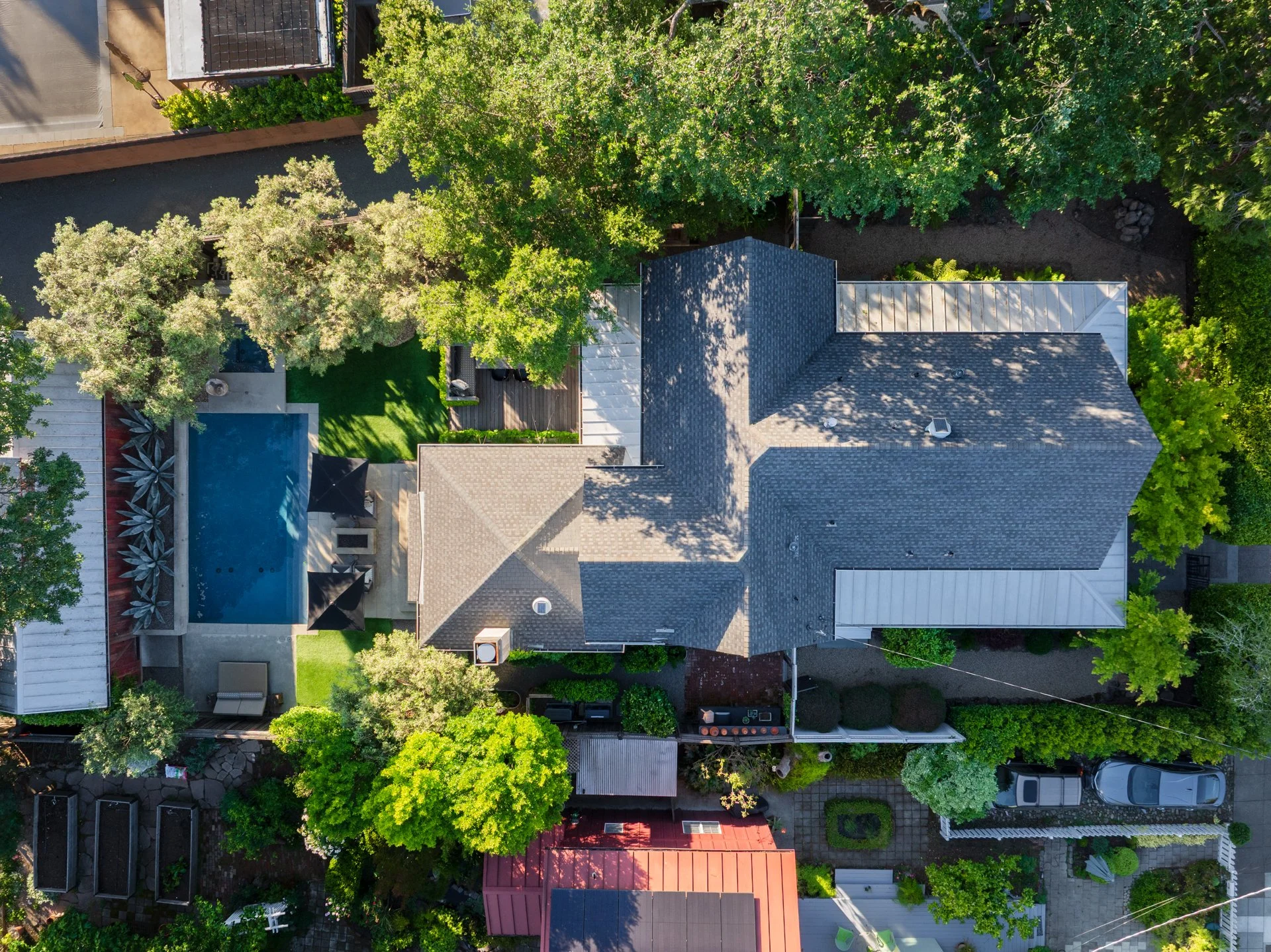 An aerial view of a residential backyard with a swimming pool, trees, and surrounding houses.