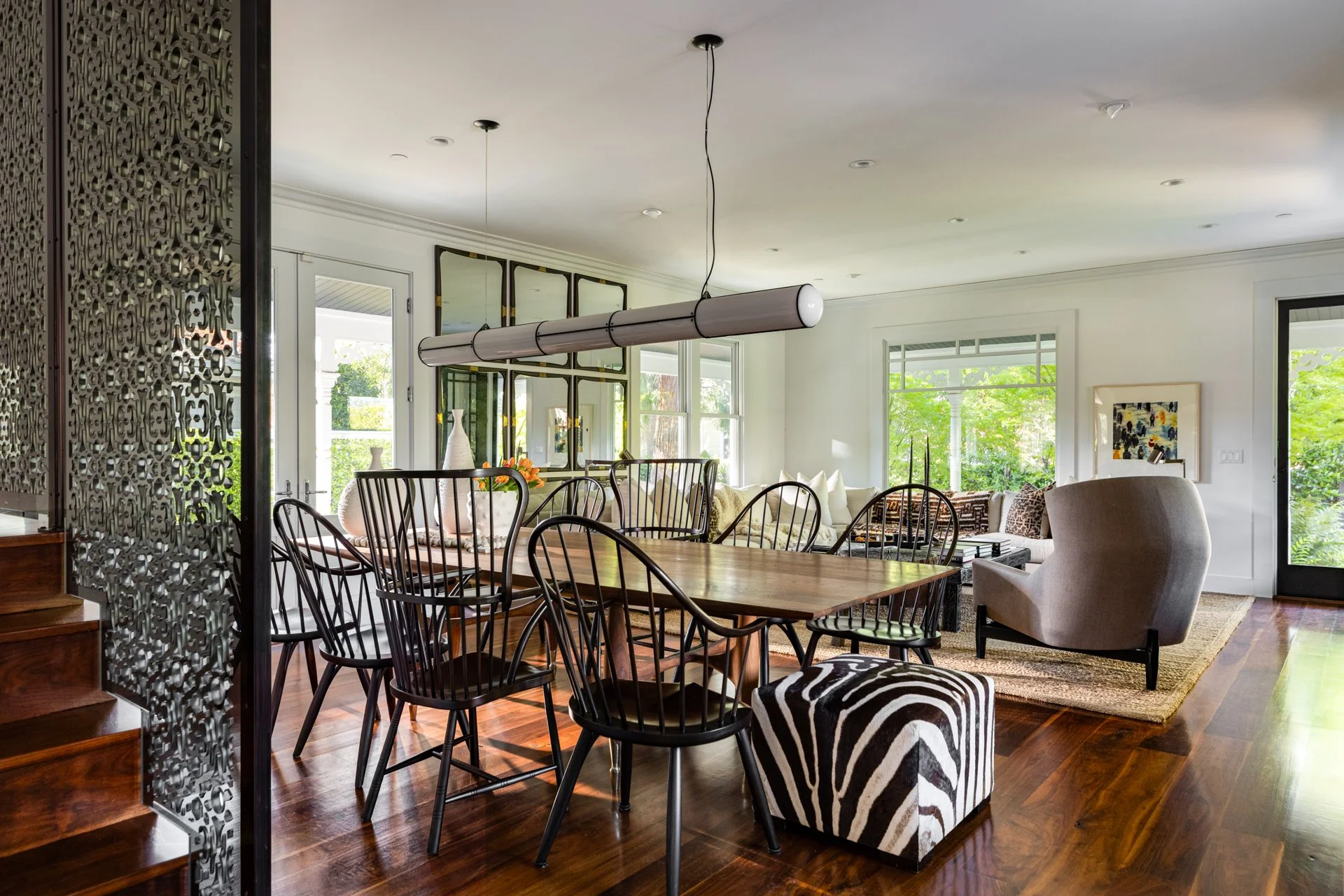 A spacious living room and dining area with large windows, modern artwork, various furniture including chairs and sofas, and decorative vases, illuminated by natural light.