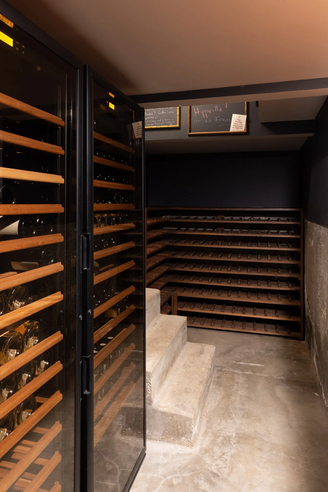 Empty wine cellar with wooden shelves, concrete steps, and a glass-door wine refrigerator.
