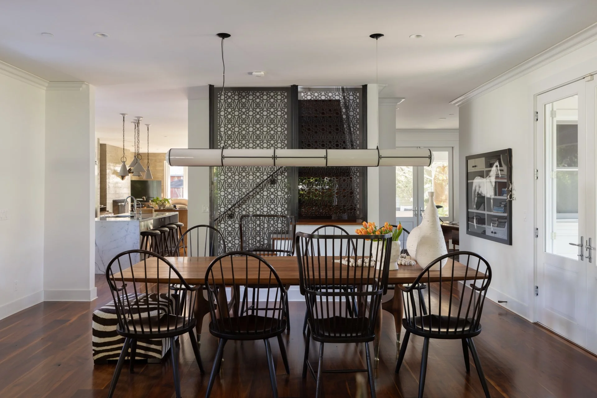 Dining area with a wooden table surrounded by black chairs, decorated with large vases and tulips, with a kitchen in the background and windows letting in natural light.