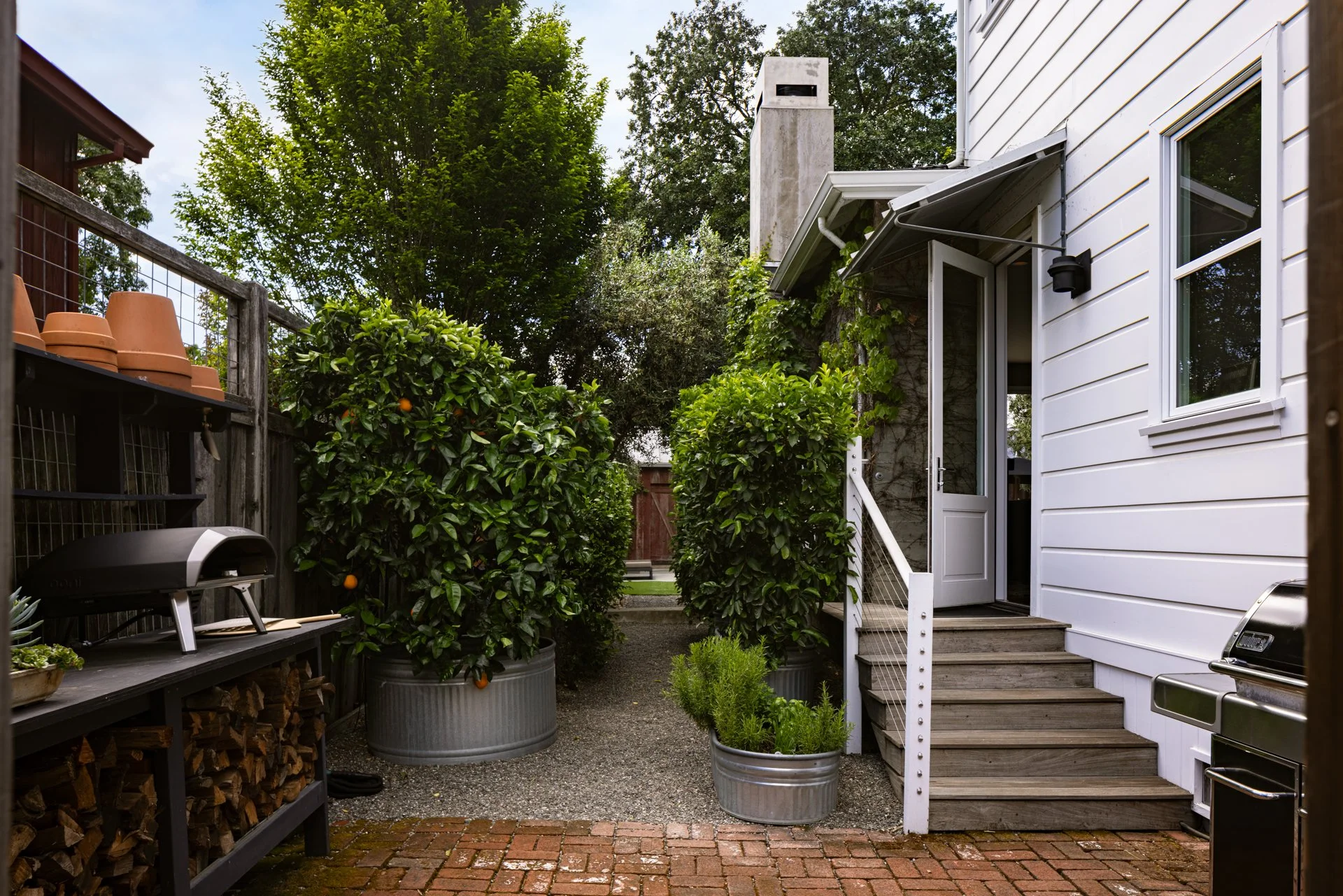 Backyard with potted trees, a wooden fence, and a white house with steps leading to a door.