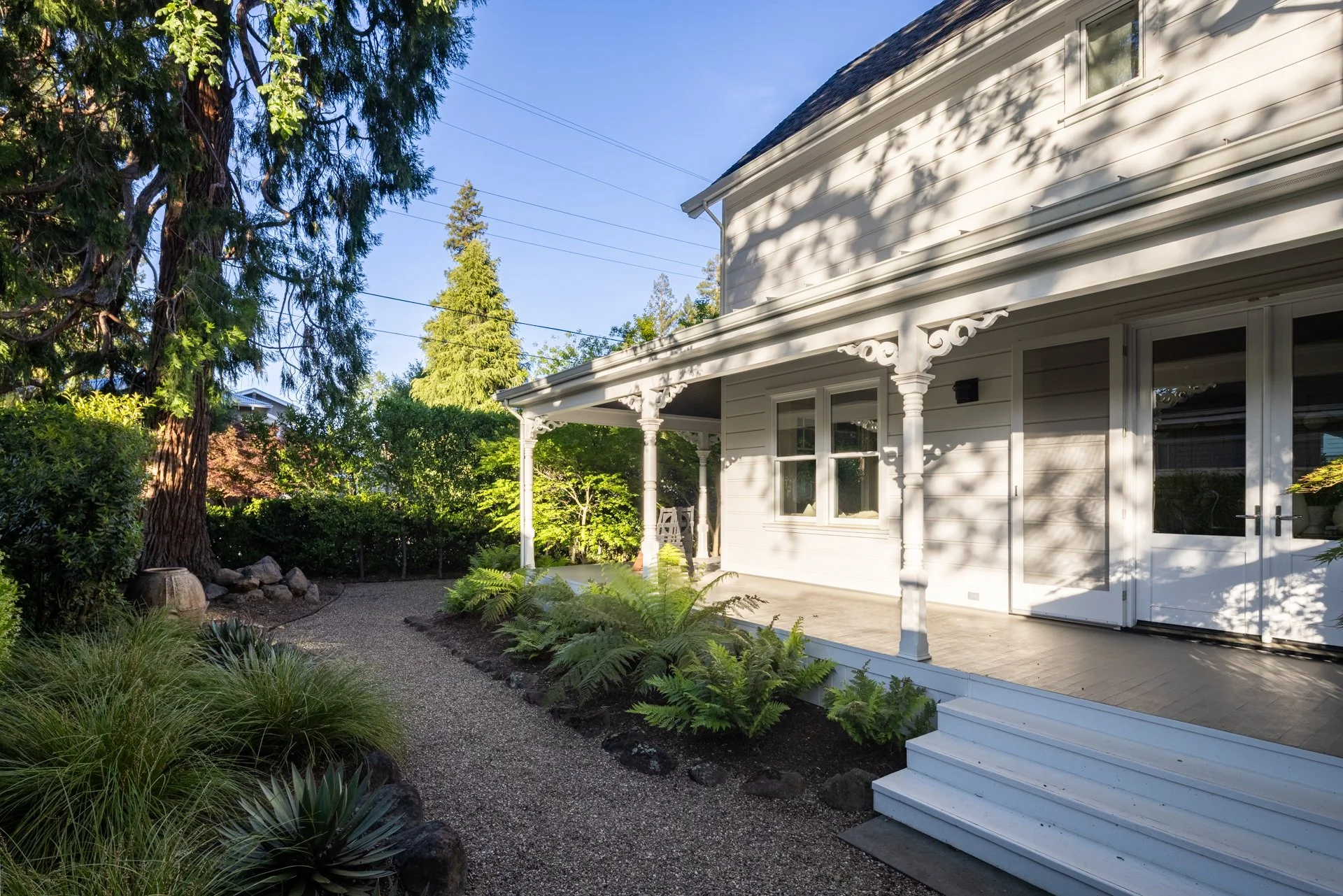 A white house with a small porch, multiple windows, and decorative columns. The porch has steps leading up to it. The yard has lush green plants, trees, and a gravel pathway, with a sunny sky overhead.