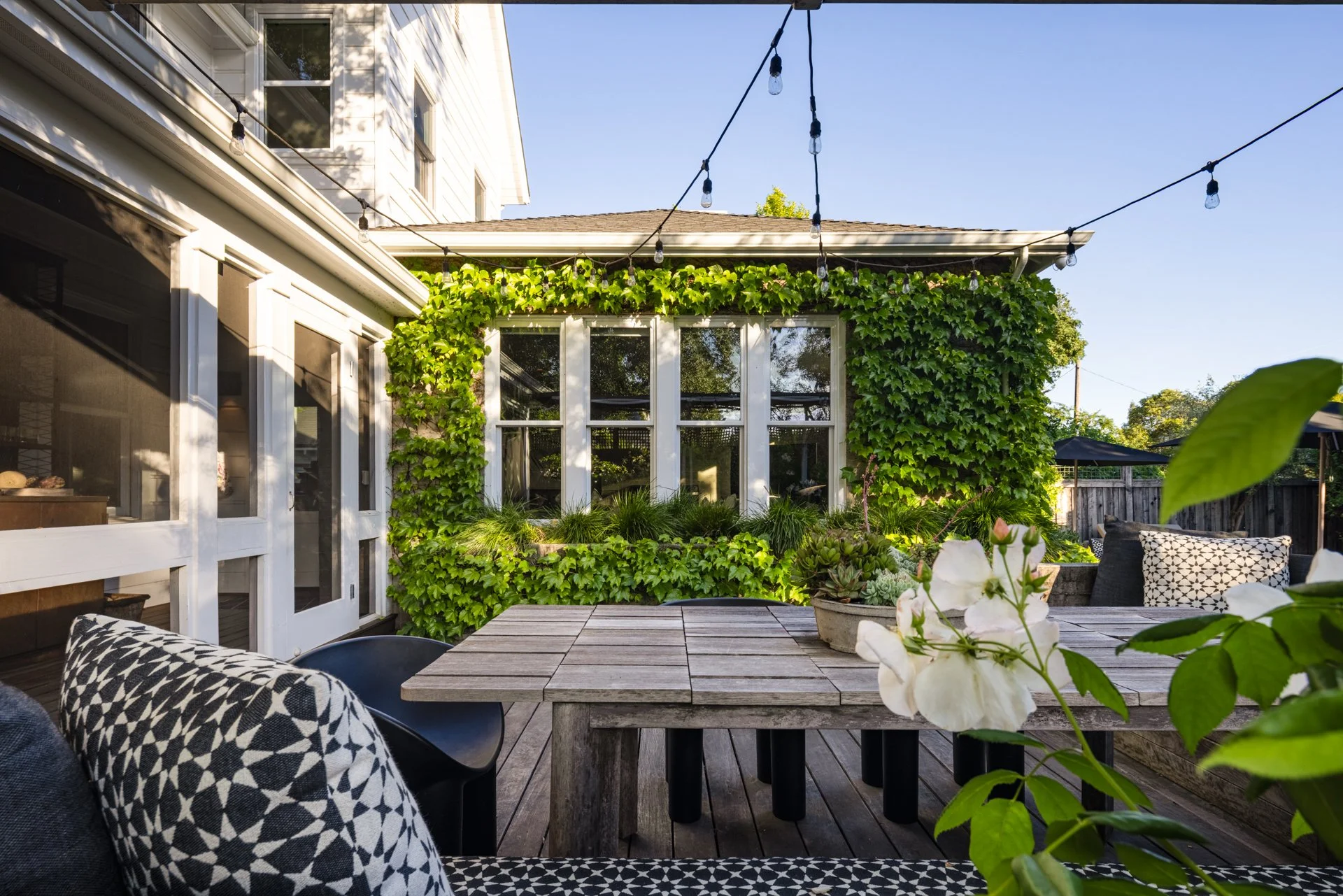 Backyard patio with string lights, lush green vines on the house, wooden table with white flowers, and outdoor seating with patterned cushions under a clear blue sky.