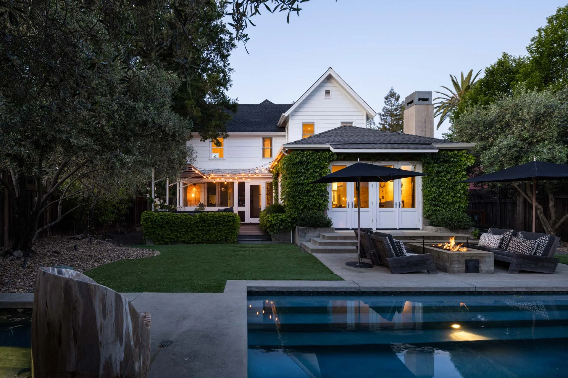 A backyard at dusk with a swimming pool in the foreground, Adirondack chairs surrounding a firepit, string lights hanging from a house, and trees and bushes surrounding the area.