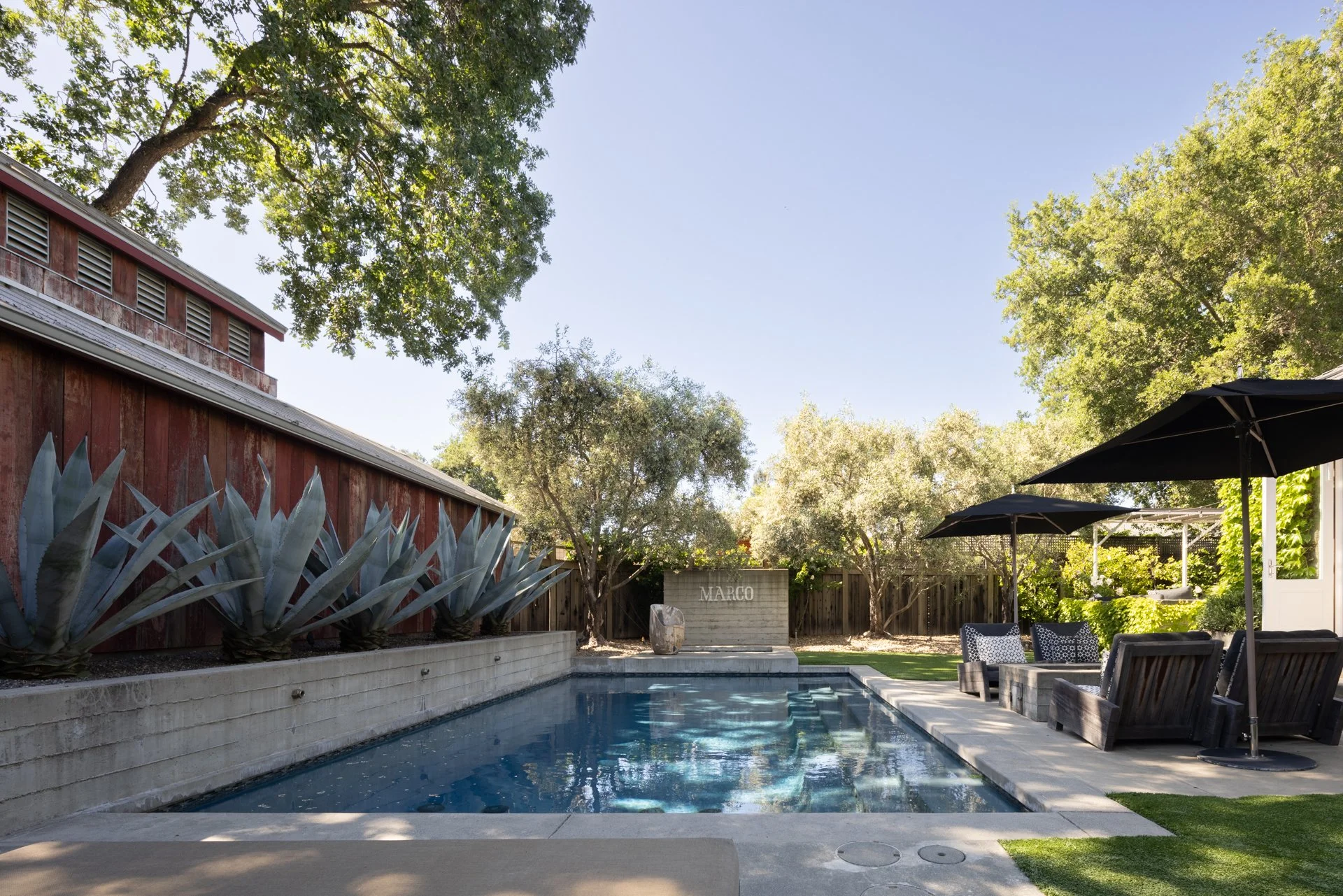 Backyard with pool, outdoor seating, large trees, umbrellas, and a building with a sign that reads 'Marco' in the background.