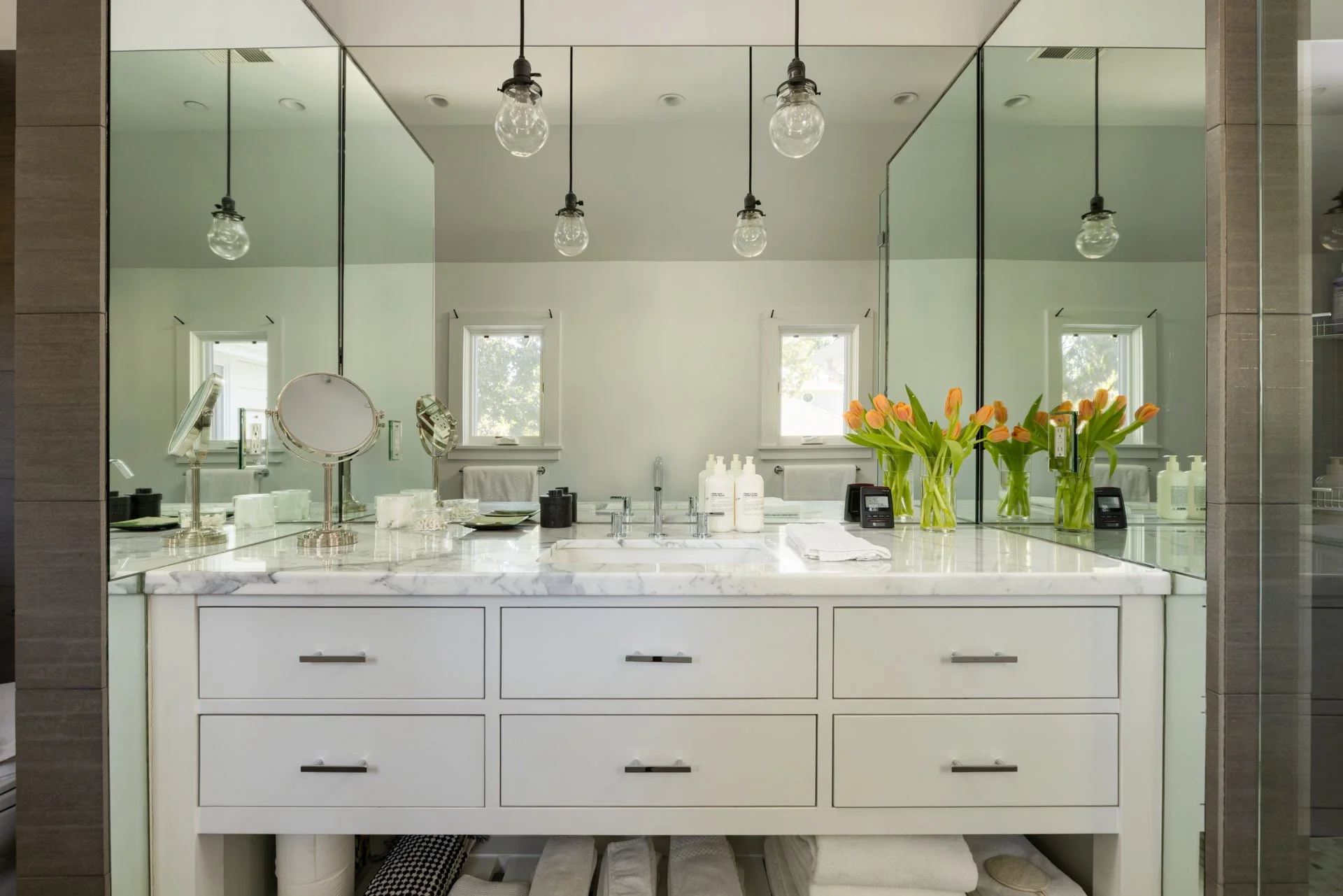 A modern bathroom vanity with a marble countertop, large mirror, two small framed windows, and decorative items including orange tulips in vases, skincare bottles, and makeup mirrors.