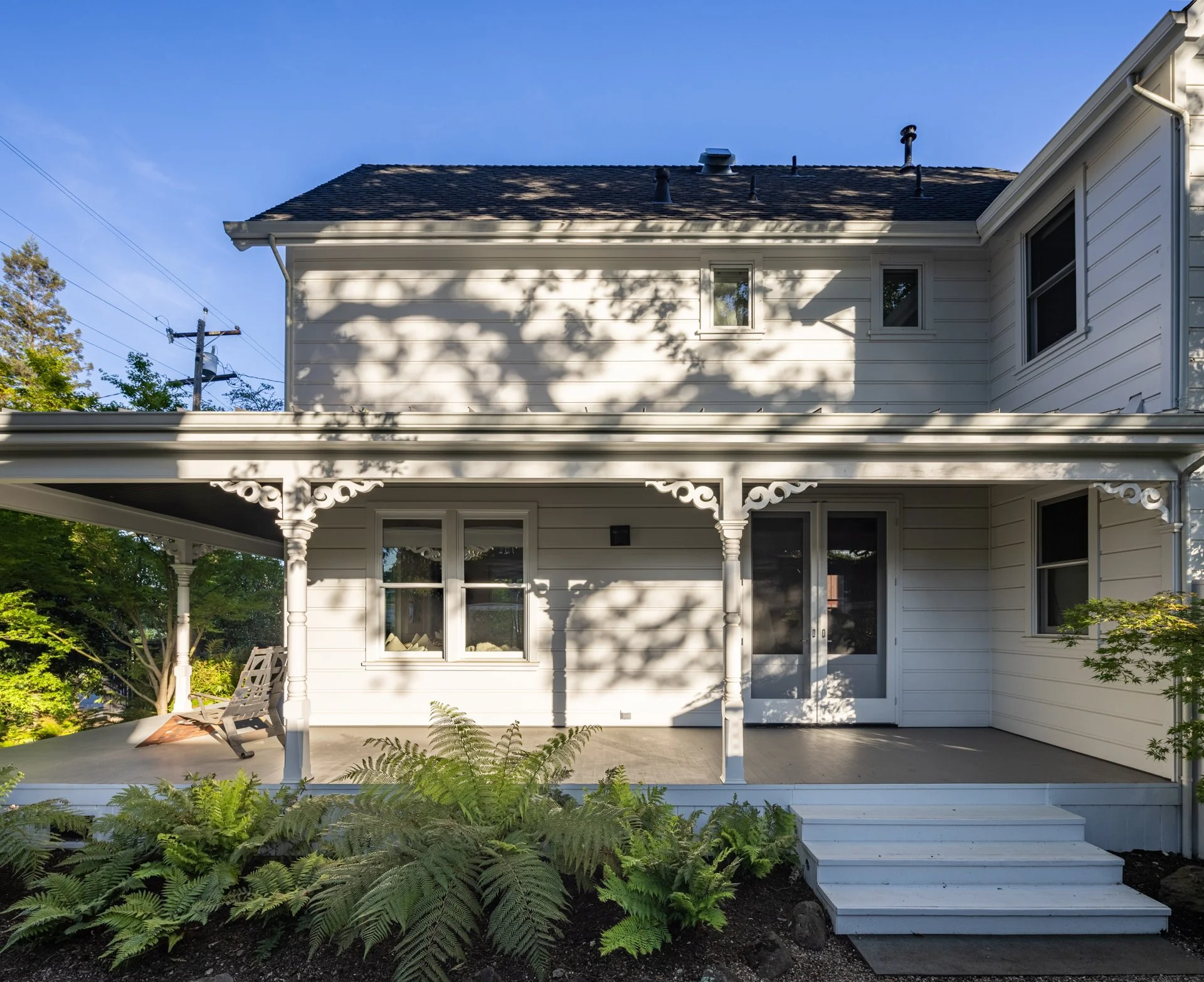 The back view of a two-story house with white siding and a porch with decorative columns. There are multiple windows on both floors, and a concrete walkway with steps leading to the porch. A bench is on the porch, and there are green plants in the fo