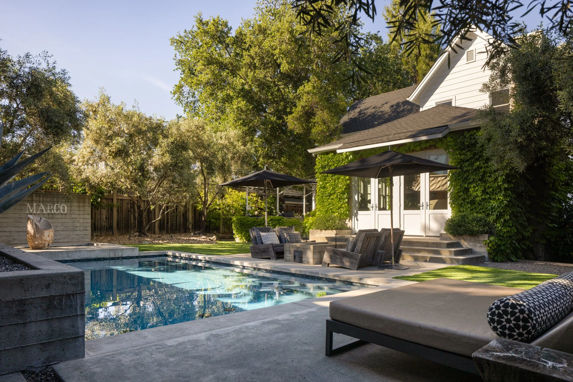 Backyard with a swimming pool, outdoor furniture including chairs and umbrellas, a white house partially covered in green ivy, surrounded by trees and a wooden fence, on a bright sunny day.