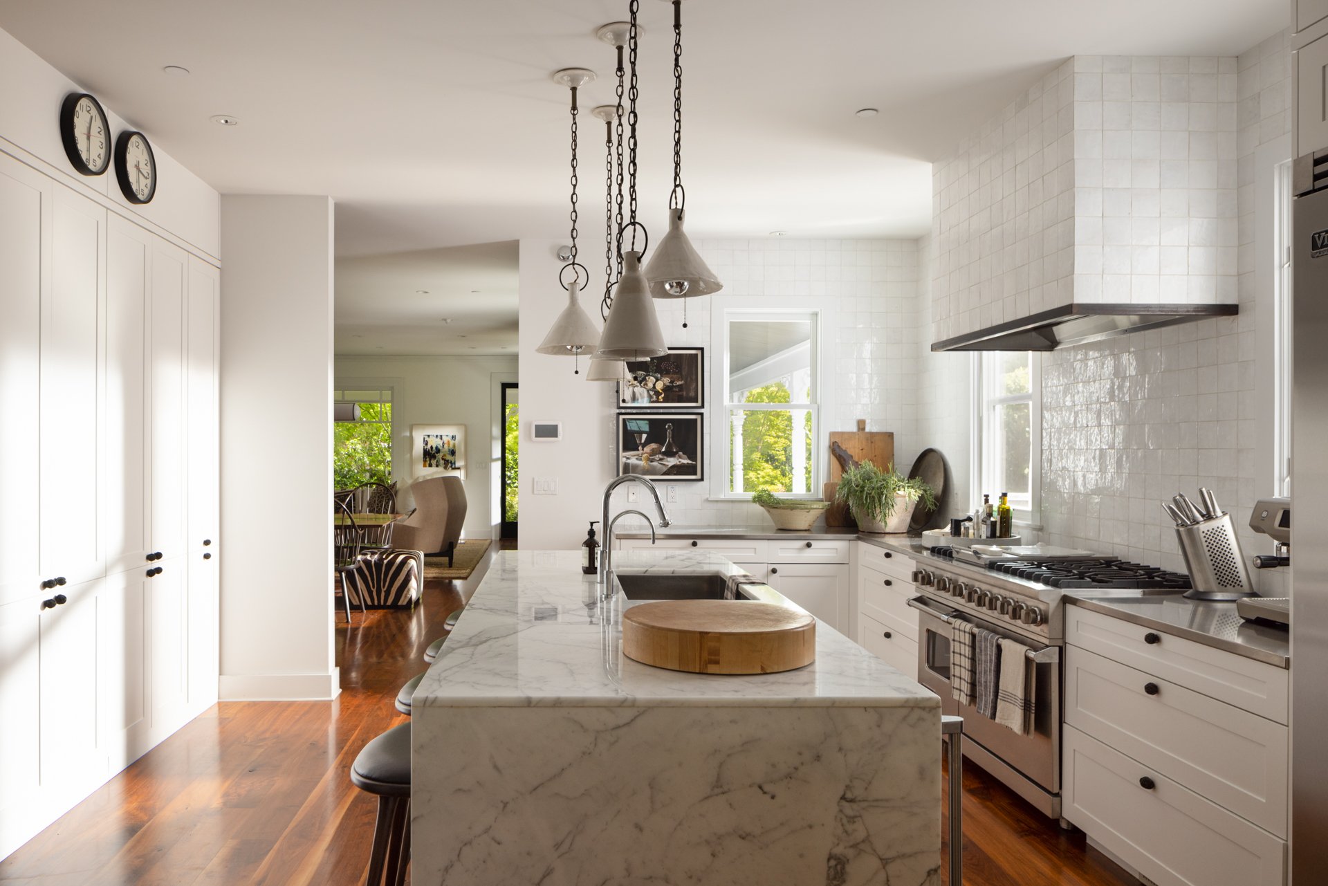 Modern white kitchen with marble island, pendant lights, and stainless steel appliances, adjacent to a dining area with chairs and artwork, natural light from windows.