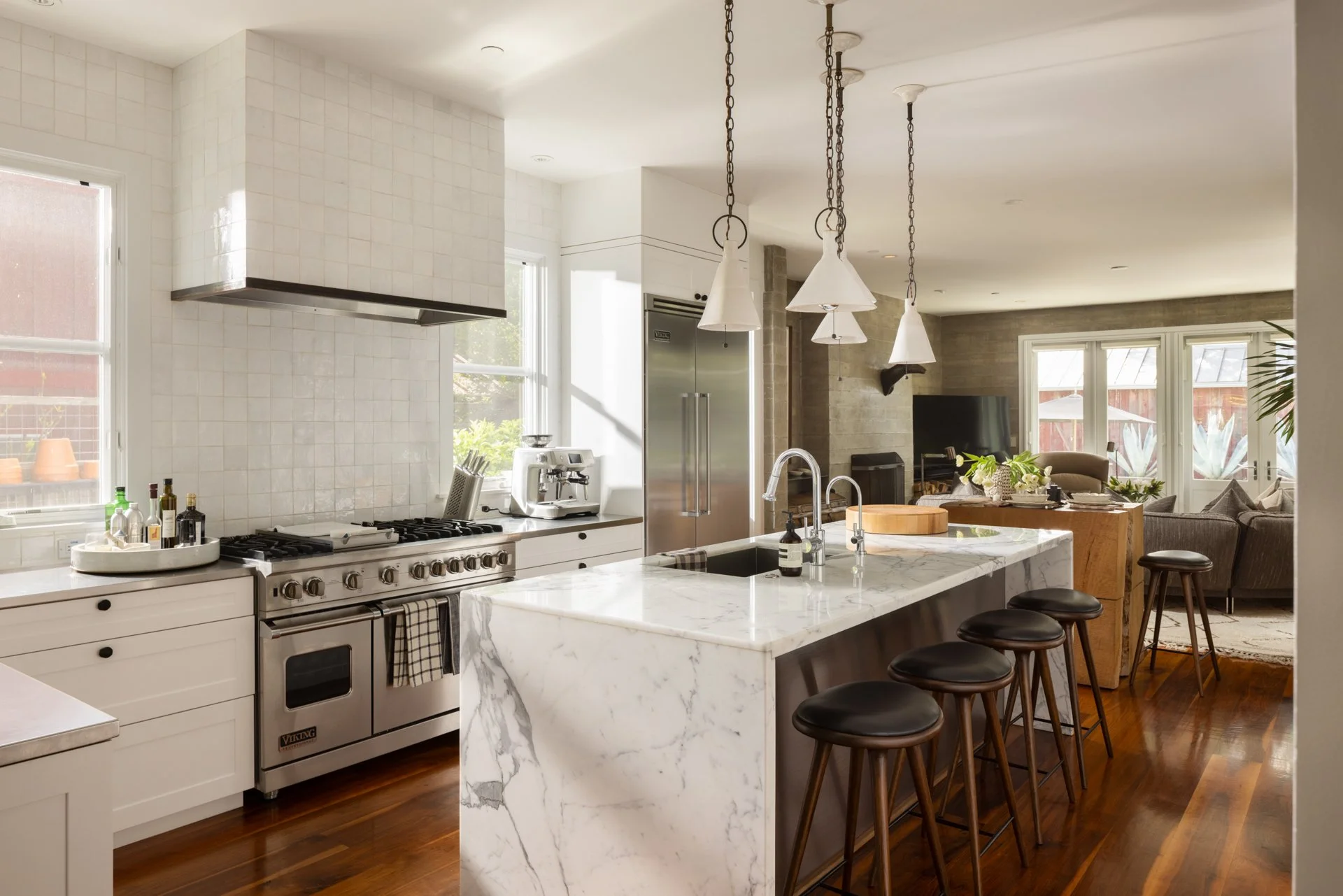 Modern kitchen with white cabinetry, marble island, stainless steel appliances, hanging pendant lights, and a view into the living room with large windows and outdoor space.