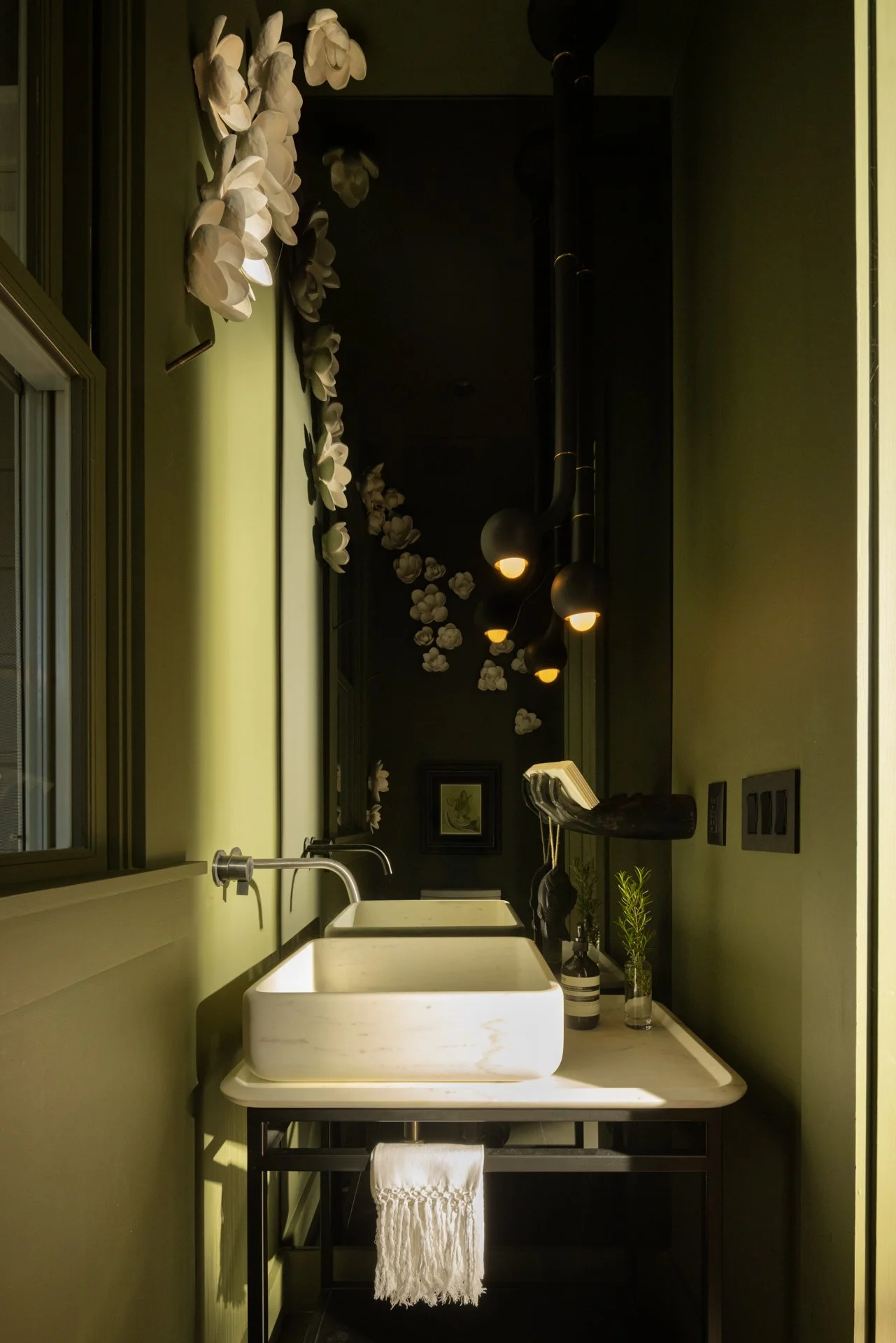 A modern kitchen sink area with a white vessel sink, a black metal frame, a white towel hanging, small potted plants, soap dispenser, green textured wall, flowers on the ceiling, black pendant lights, and framed artwork in the background.