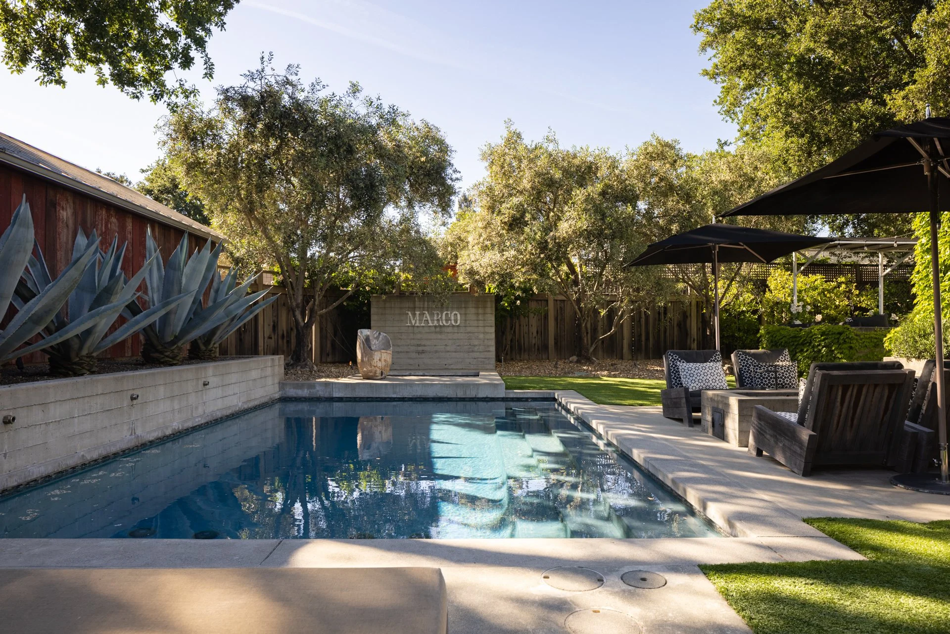A backyard with a swimming pool, a wooden fence with trees, outdoor furniture under umbrellas, and a stone sign with the name 'MARCO' on it.