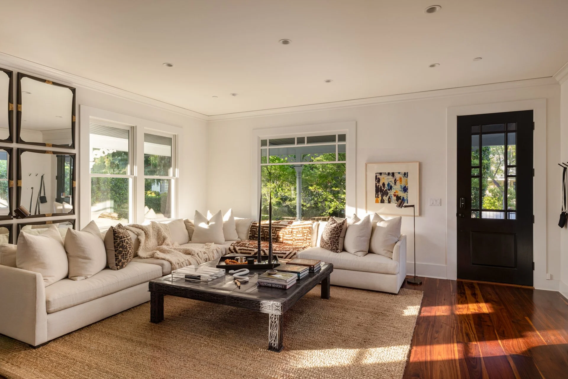 A bright living room with white walls, large windows, a white sectional sofa with various pillows, a black coffee table with books and decor, and a black front door with glass panels. There's artwork on the wall and hardwood flooring.