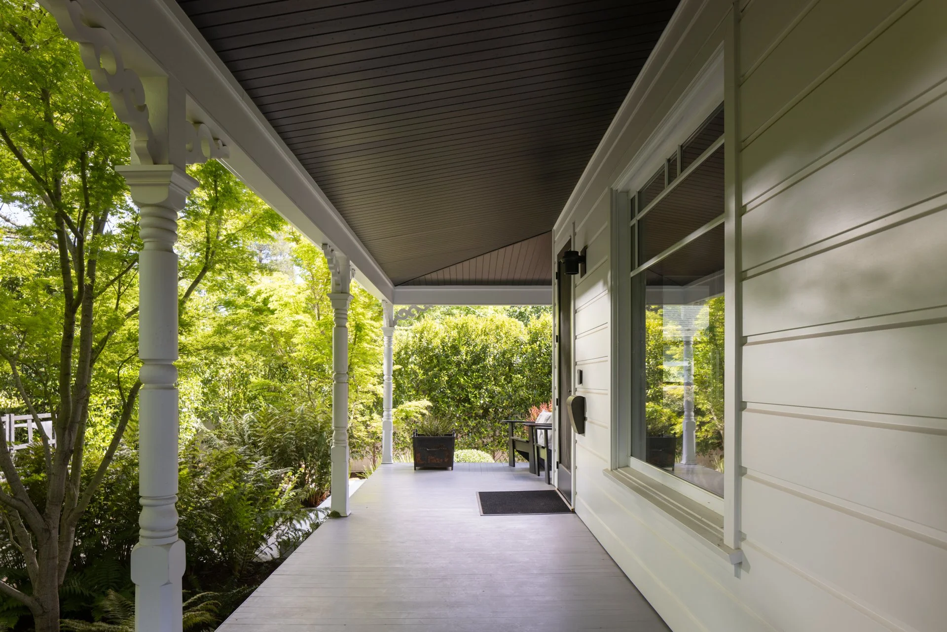 A porch with white painted wood flooring, decorative white columns, beige siding, and a dark brown ceiling. Green trees and bushes are visible in the background.
