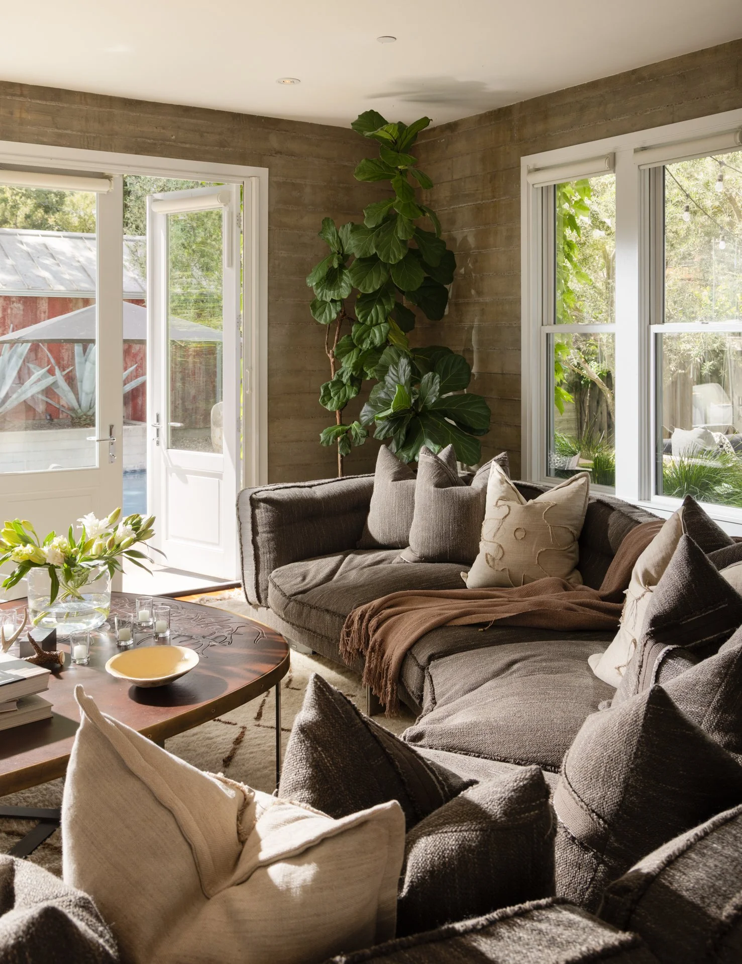 Living room with a gray sectional sofa, beige and cream pillows, a brown throw blanket, a wooden coffee table with candles, a vase of white flowers, large windows showing greenery outside, and a tall indoor fig tree near the corner.