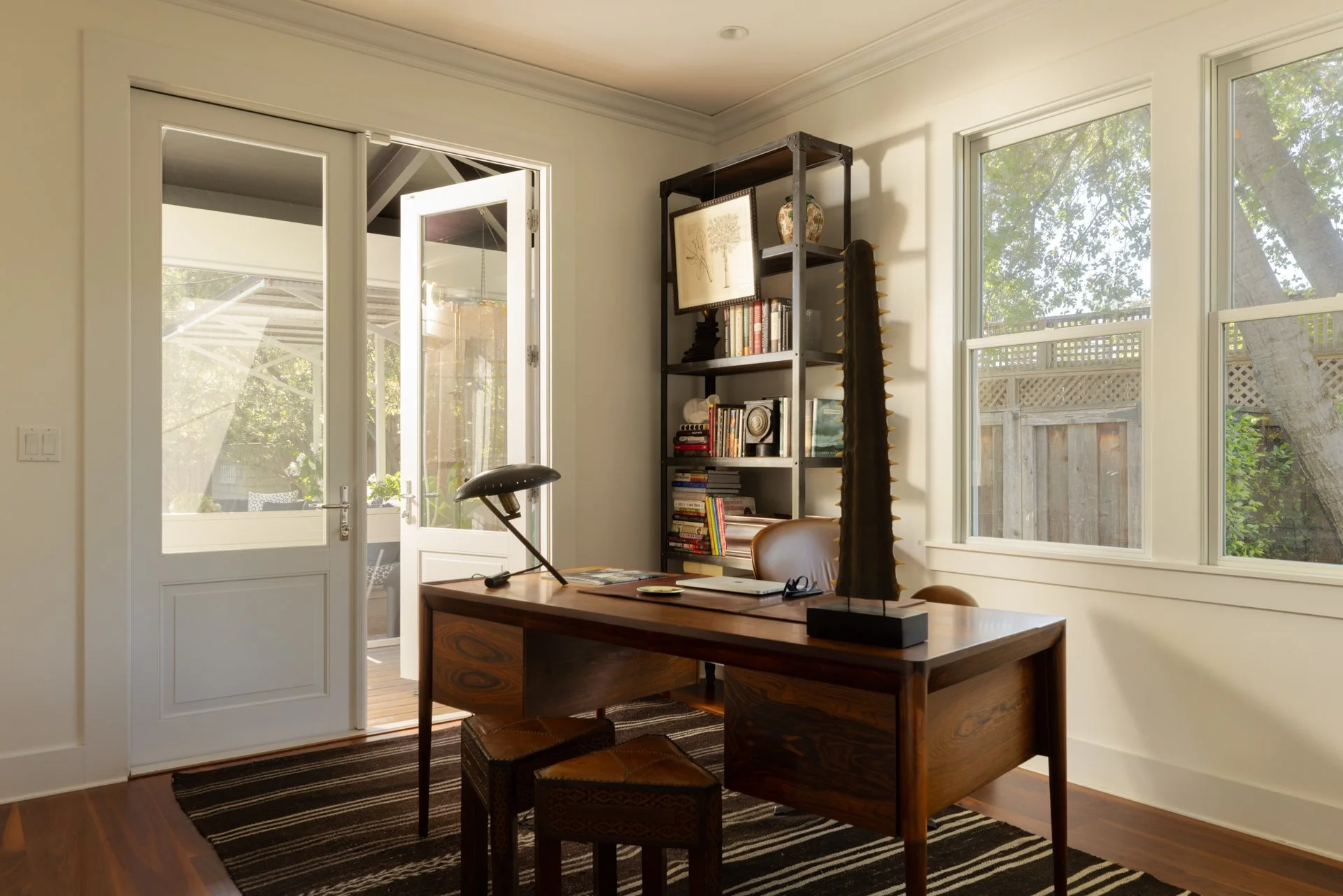 A home office with a wooden desk, two stools, a black bookshelf filled with books and decorative items, and large windows showing trees outside. An open door leading to a porch is on the left.